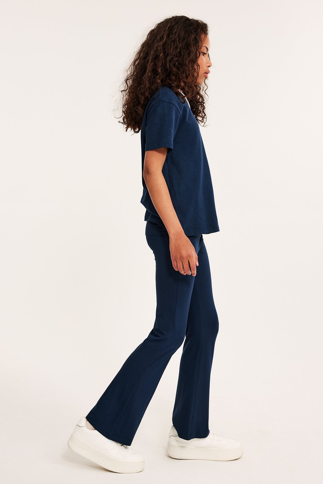 Widok z boku dziecka w niebieskich rozszerzanych legginsach do jogi, niebieskiej koszulce i białych sneakersach.