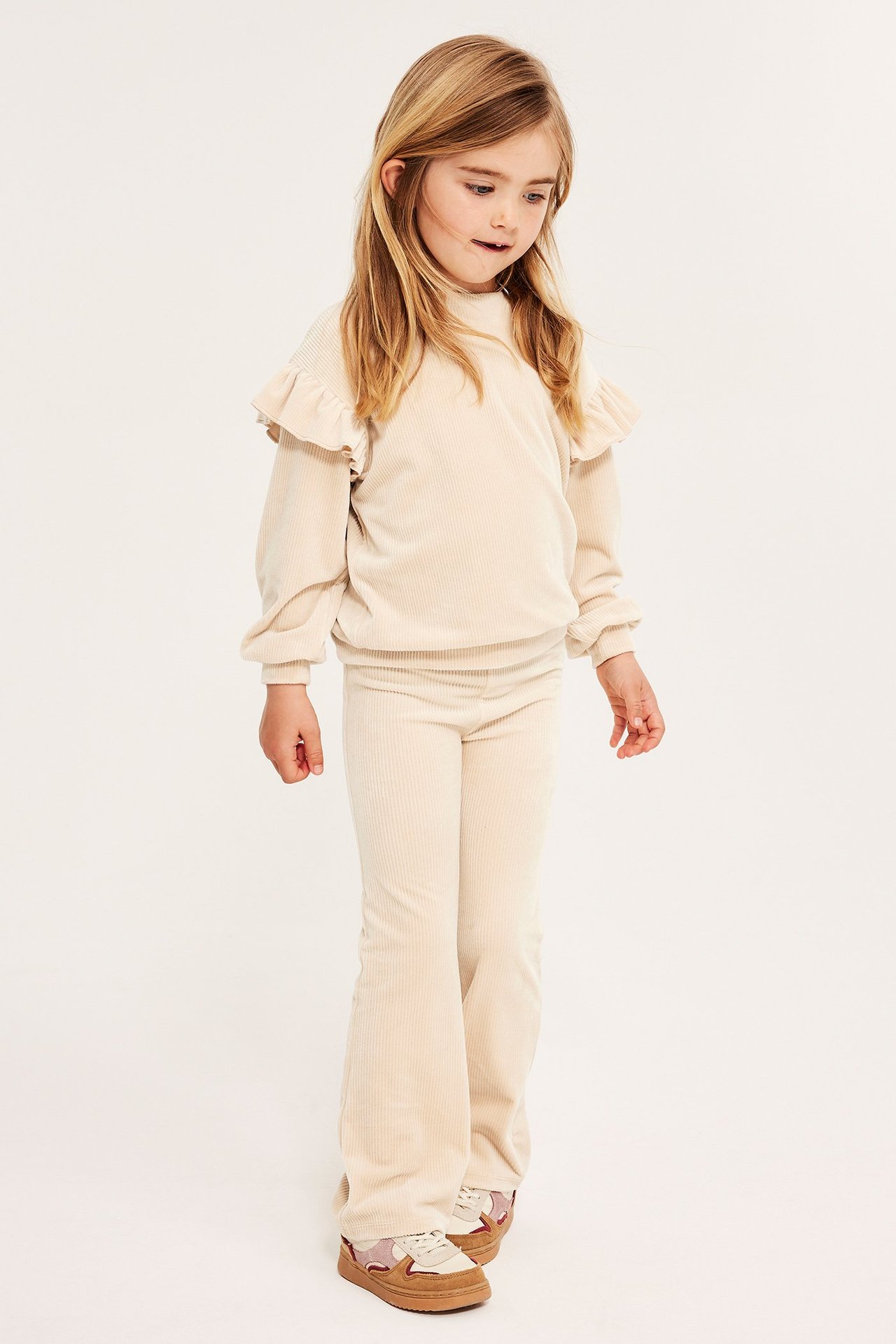 Widok z przodu dziecka w beżowych prążkowanych welurowych legginsach, pasującym topie z falbankami i sneakersach.