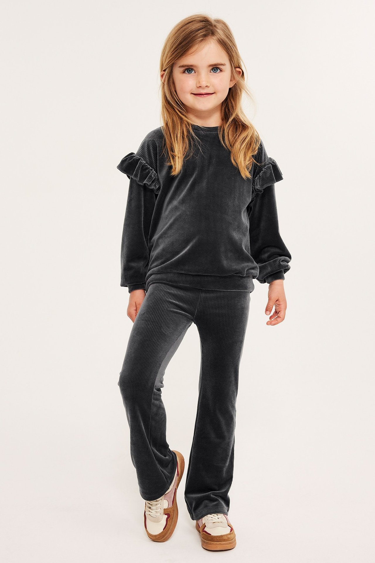 Widok z przodu dziecka w ciemnoszarych prążkowanych welurowych legginsach dzwonach, pasującym topie z falbankami i sneakersach.