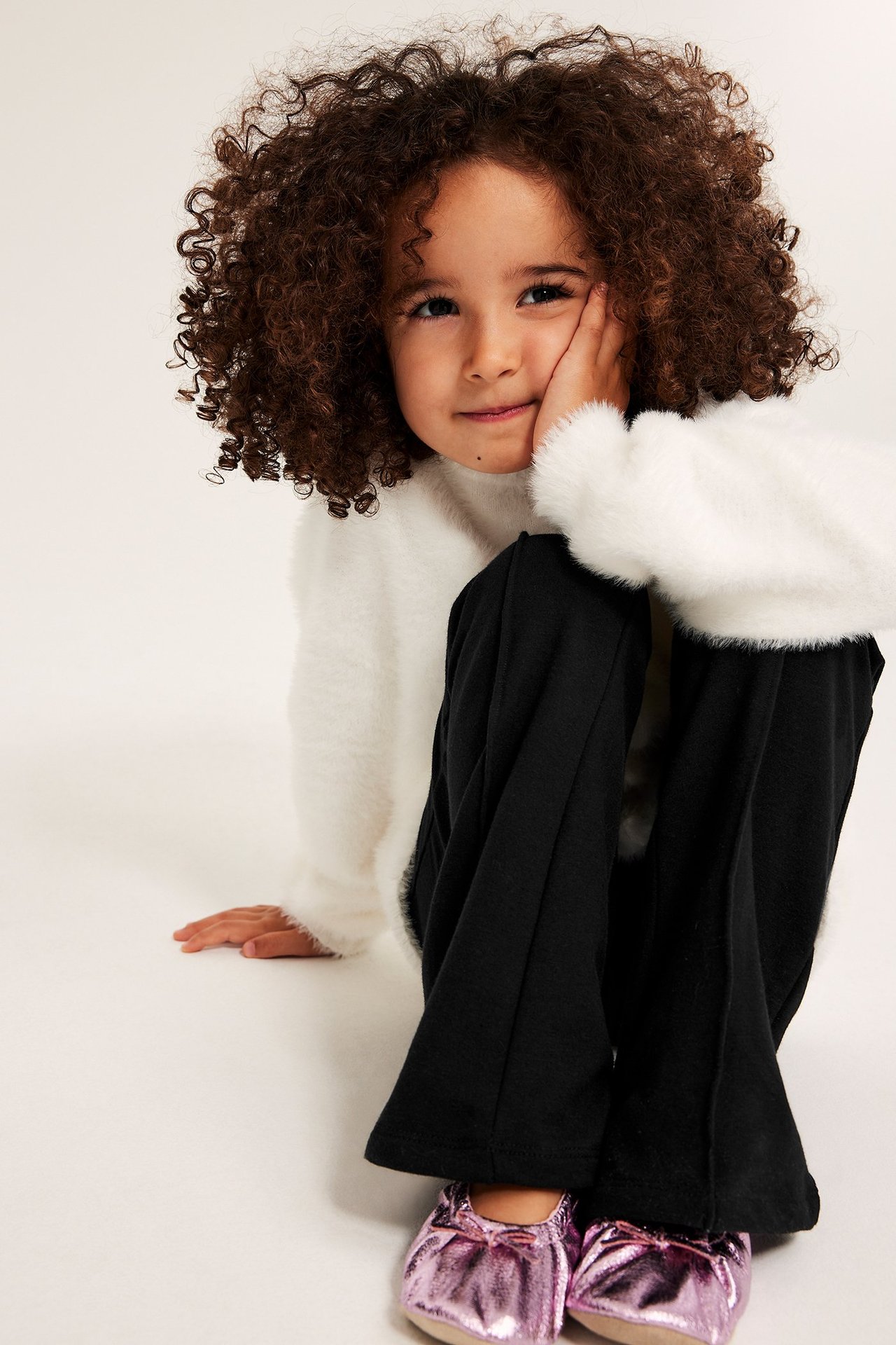 Edestä: lapsi mustissa levenevissä trikoohousuissa, valkoisessa pörröneuleessa ja kiiltävissä vaaleanpunaisissa ballerinoissa.