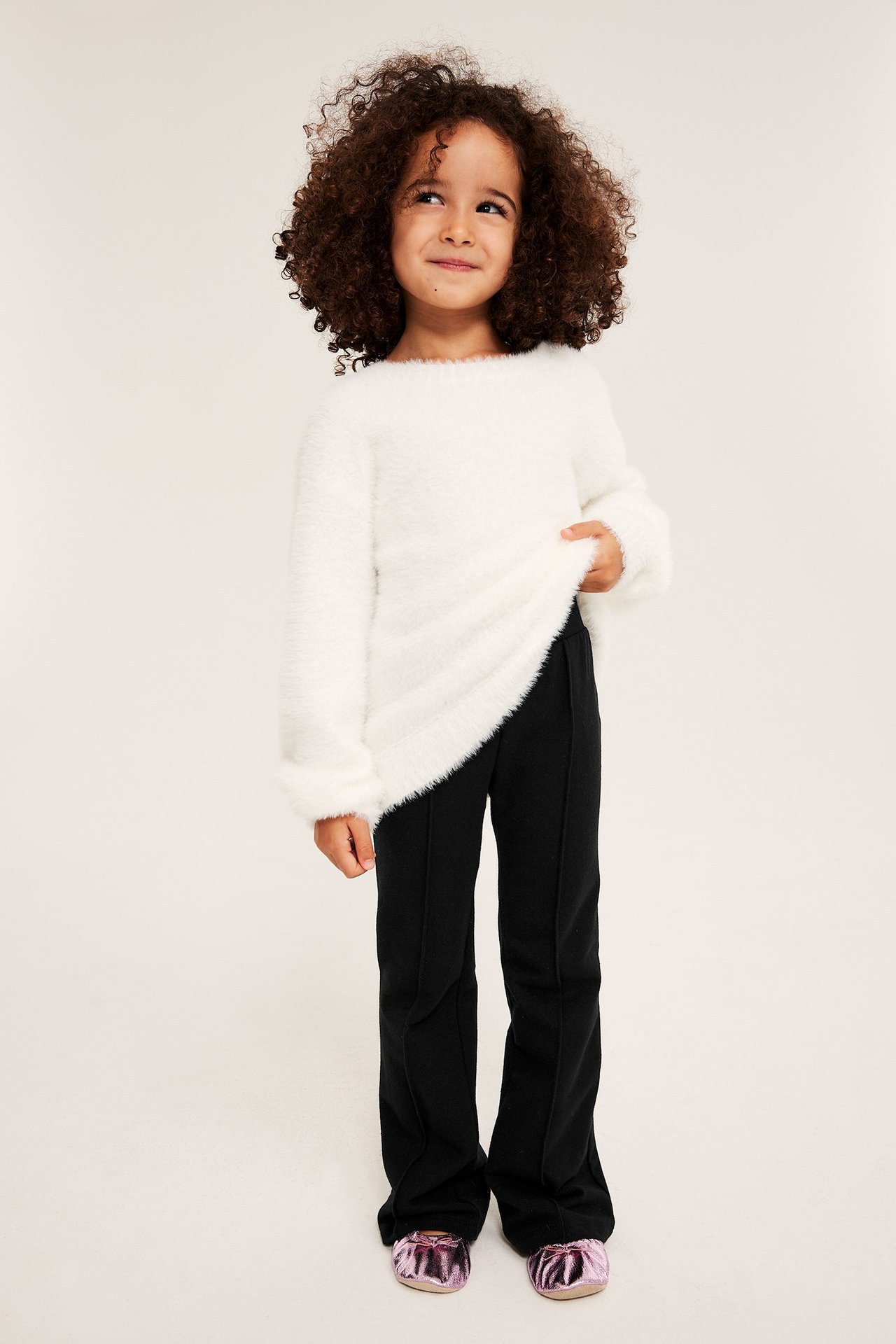 Edestäpäin: lapsi mustissa levenevissä trikoohousuissa, valkoisessa pörröneuleessa ja pinkeissä metallikengissä.