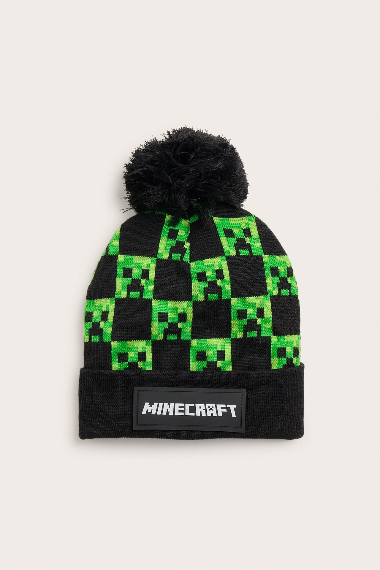 Framifrån: svart stickad barnmössa med grönt Minecraft Creeper-mönster och svart tofs.
