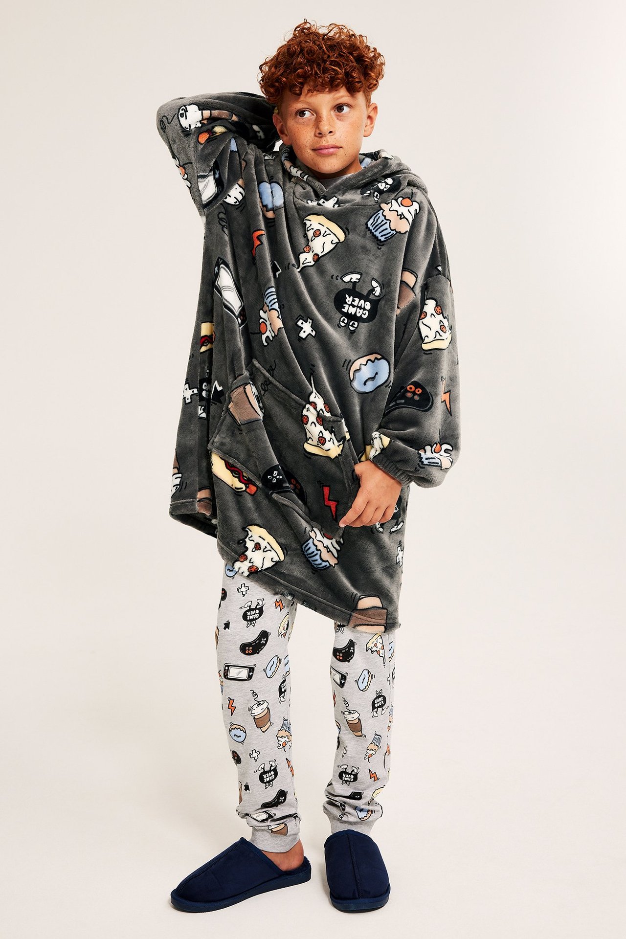 Widok z przodu: Dziecko w za dużej, ciemnoszarej, wzorzystej bluzie polarowej z kapturem, jasnoszarych spodniach i granatowych kapciach.