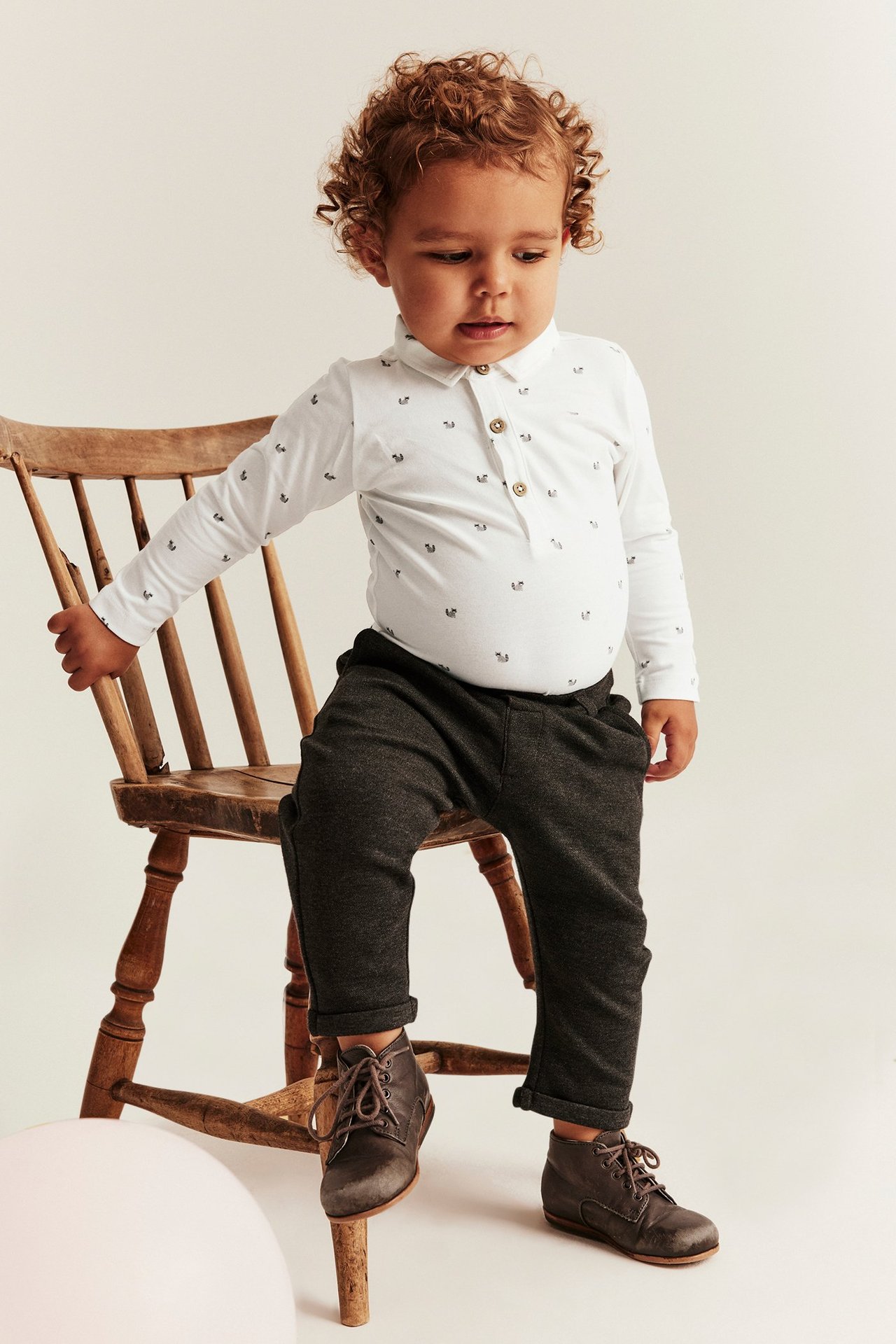 Framifrån, en baby i offwhite långärmad mönstrad body med krage, mörkgrå byxor och bruna kängor.