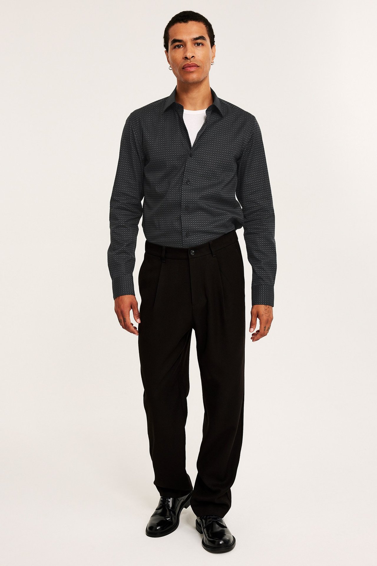 Man framifrån i mörkgrå mönstrad bomullsskjorta över vit t-shirt, svarta plisserade byxor och svarta finskor.