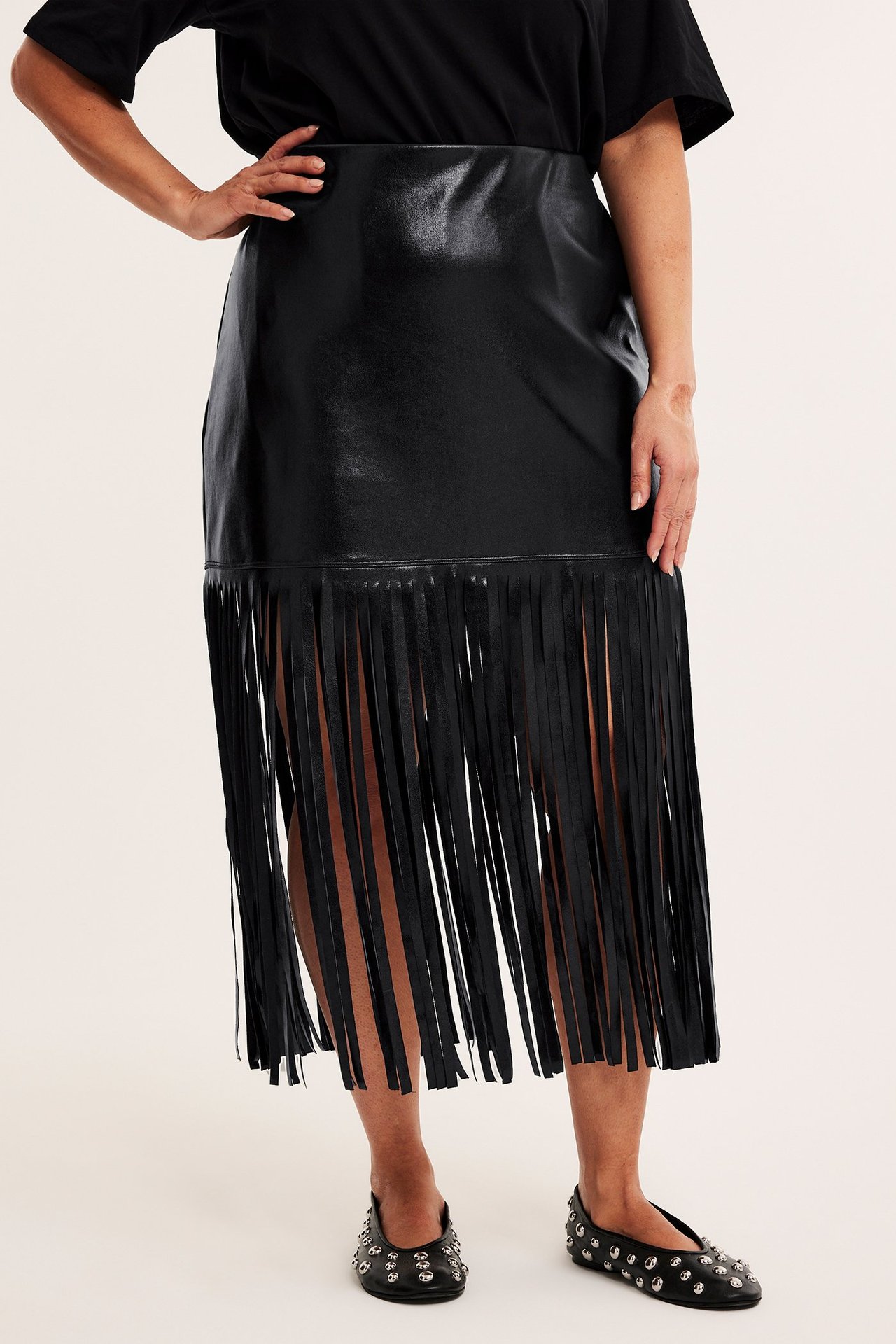 Edestä: Nainen mustassa tekonahkahameessa hapsuilla, mustassa t-paidassa ja mustissa niittibalettikengissä.