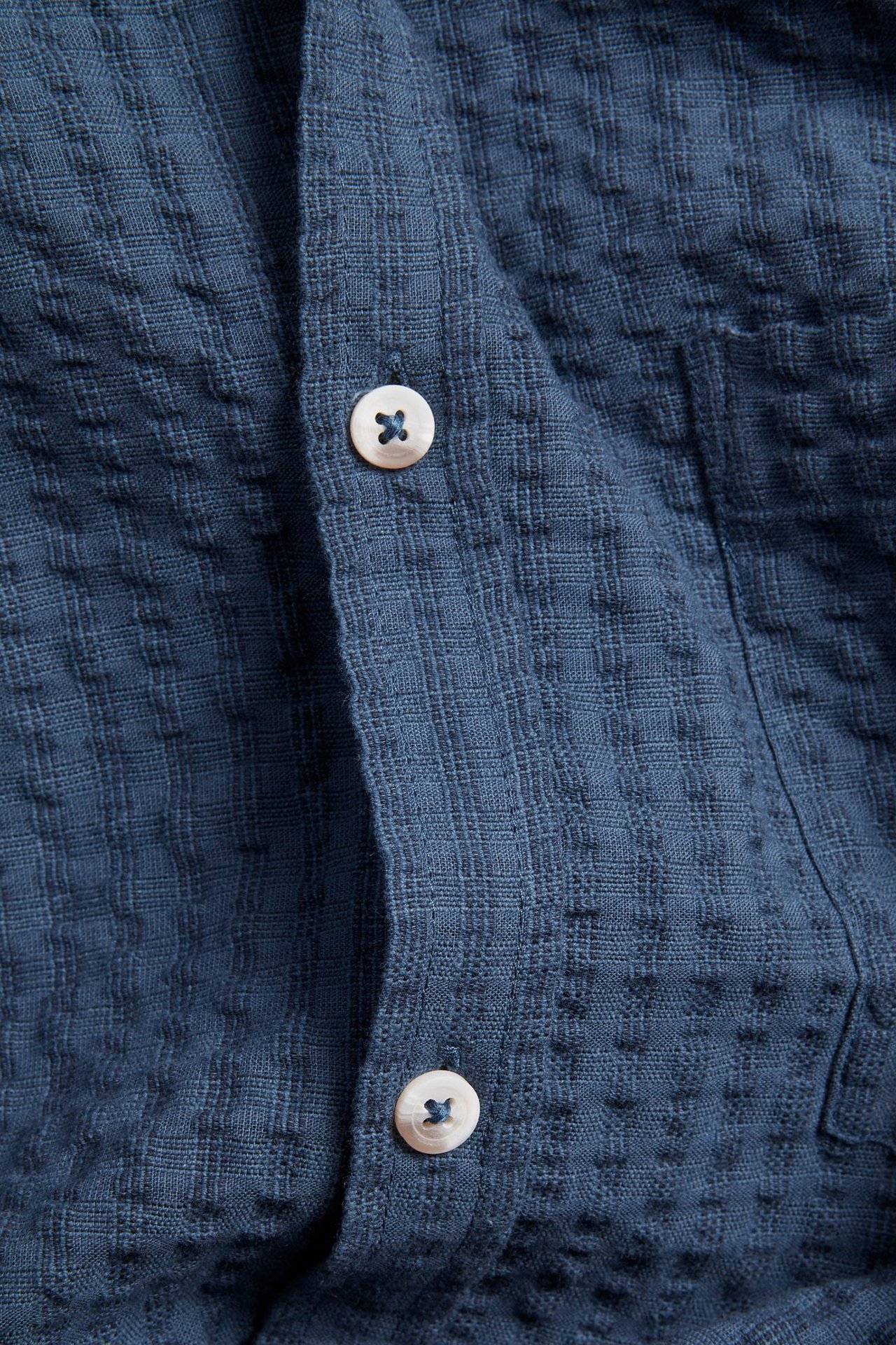 Zbliżenie ciemnoniebieskiej, strukturalnej koszuli bawełnianej z białymi guzikami na plisie.