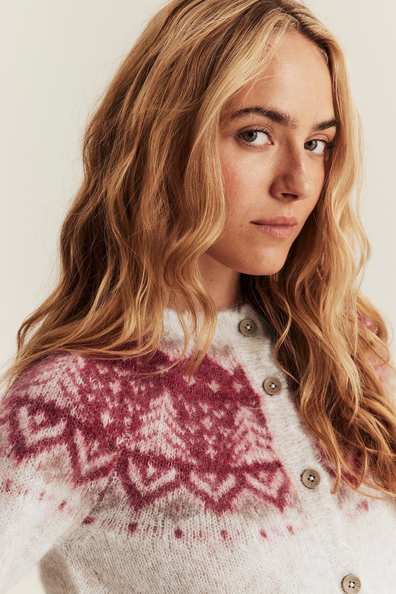 Nærbilde, forfra: Kvinne i lys beige melert ullblanding jacquard strikkejakke med røde mønstre og knapper.