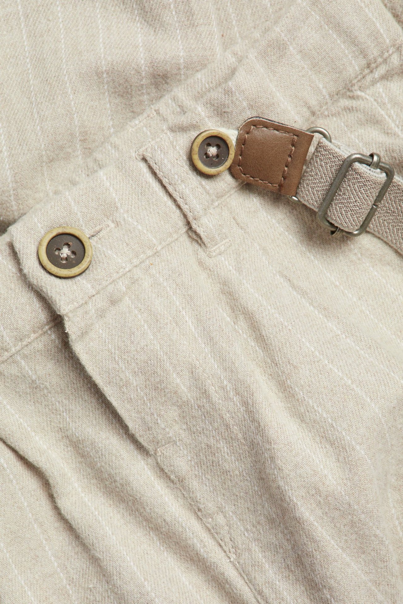 Nærbilde av beige, stripete vevde snekkerbukser med knapper, imitert skinndetalj og metallspenne.