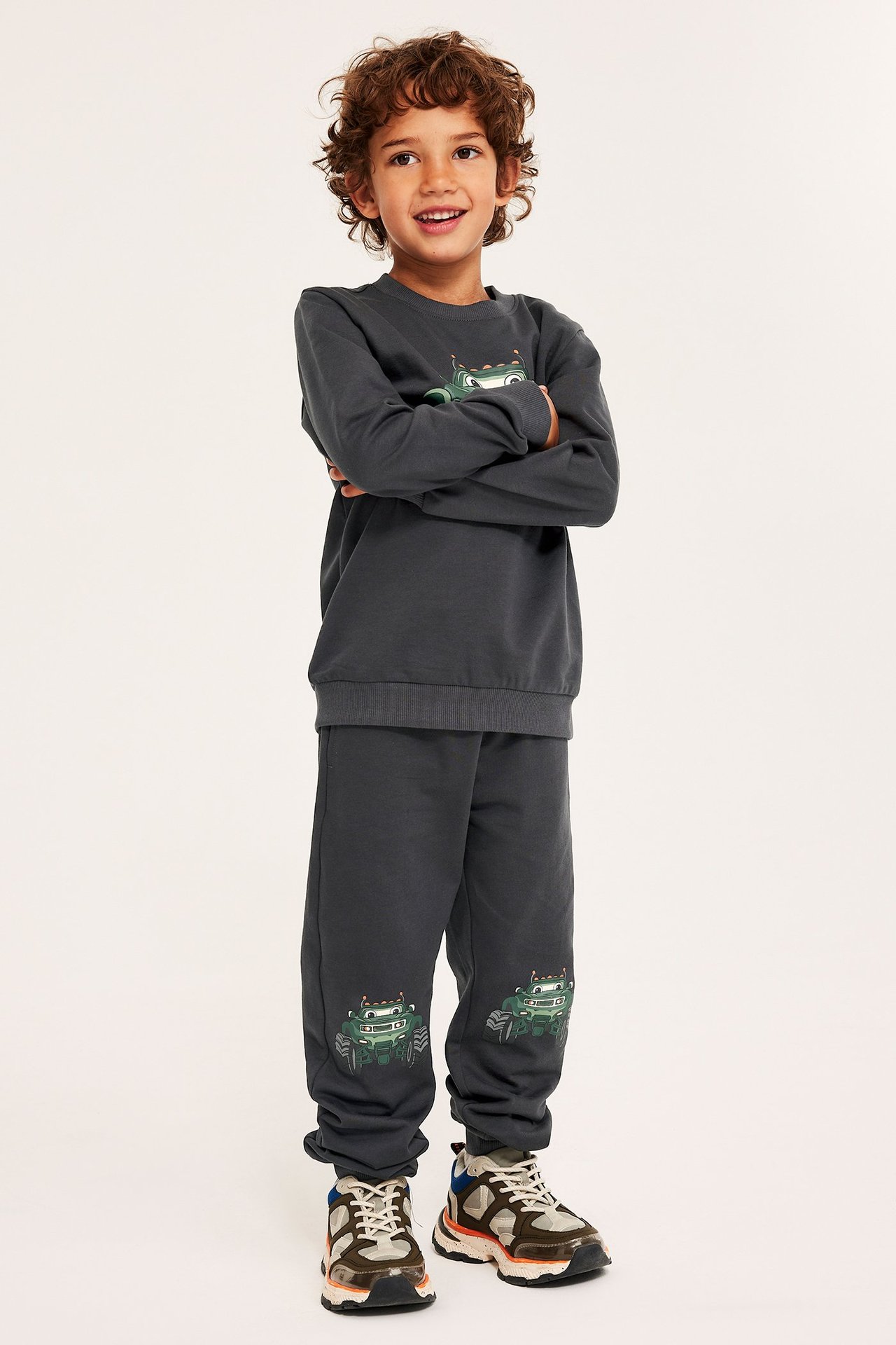 Edestä kuvattu lapsi tummanharmaissa collegehousuissa, joissa monsteriautoprintti, yhteensopiva paita ja lenkkarit.