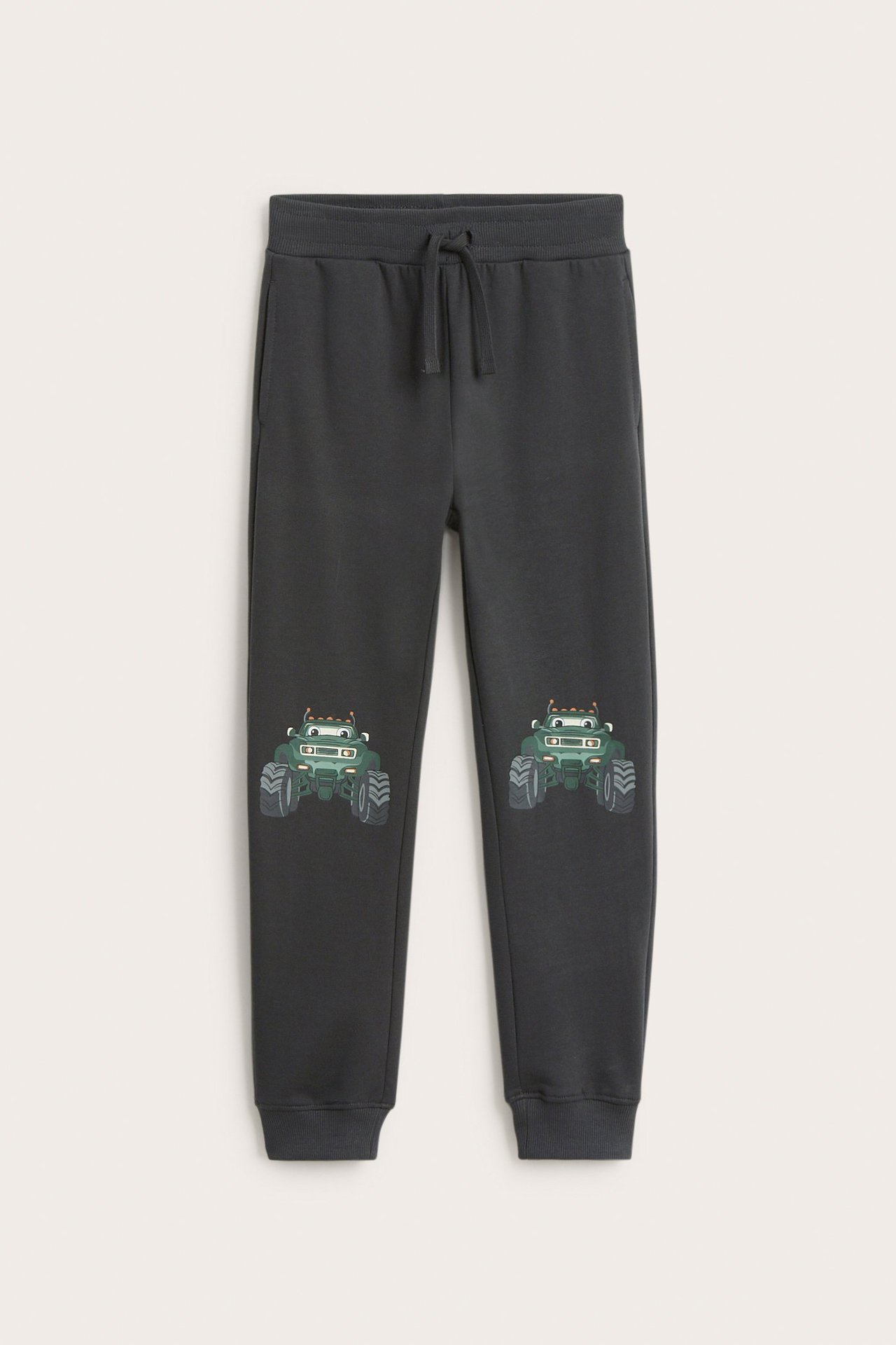 Edestä: lasten mustat puuvillaiset collegehousut, monstertruck-printit polvissa, resori vyötäröllä, kiristysnyöri.