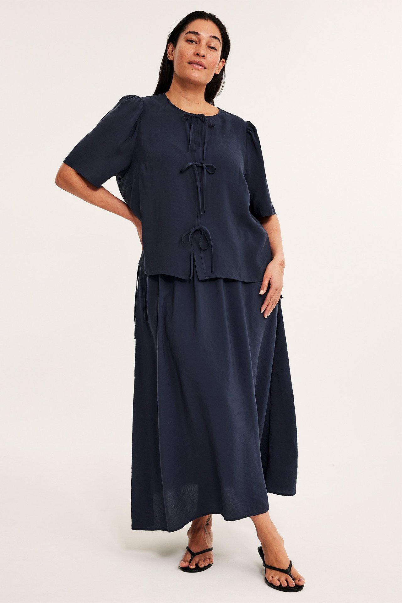 Frontvy: Kvinna i mörkblå knytblus med puffärm, matchande maxikjol och svarta sandaler.