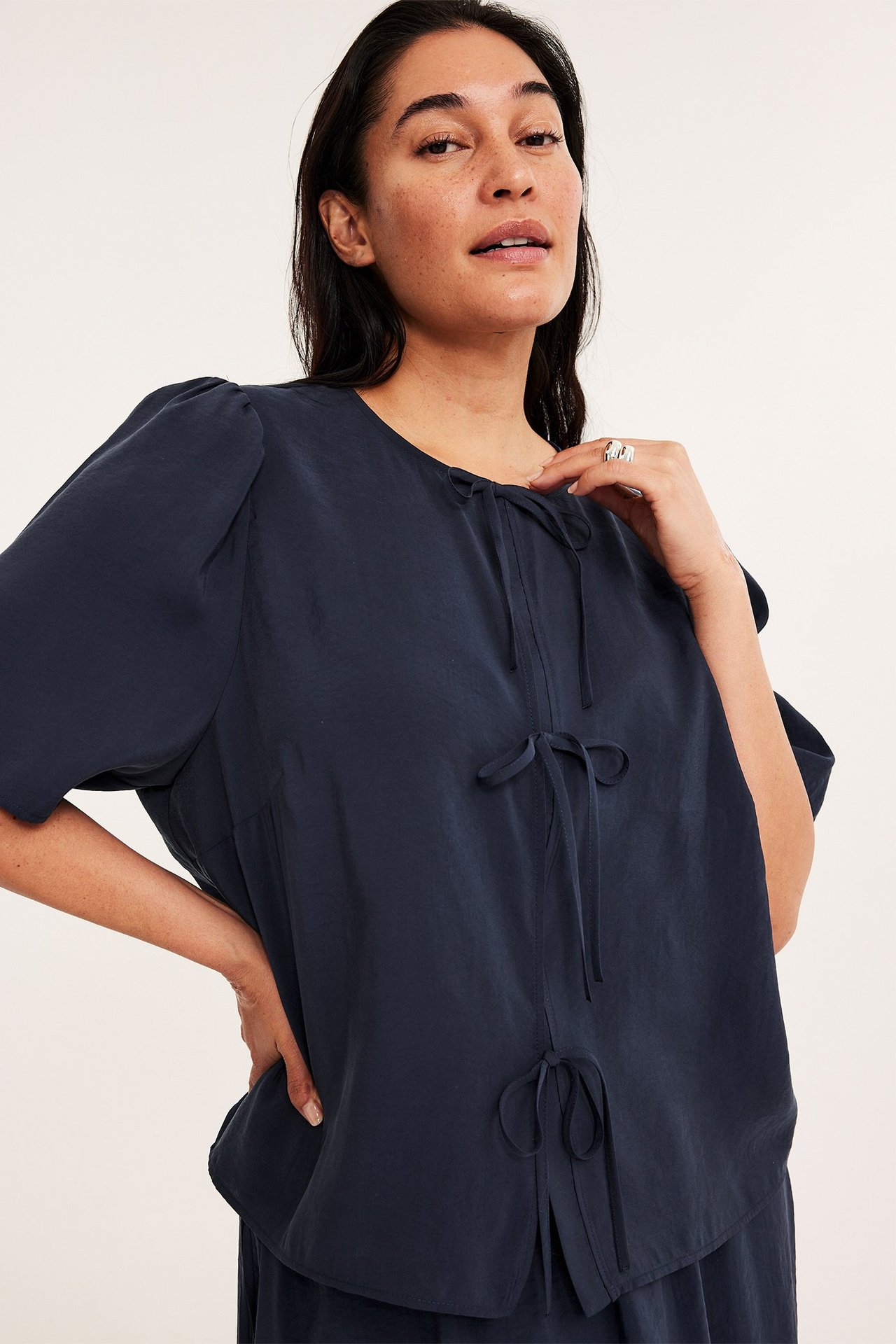 Z przodu: kobieta w granatowej bluzce wiązanej z modalu, bufiastymi rękawami, srebrny pierścionek.