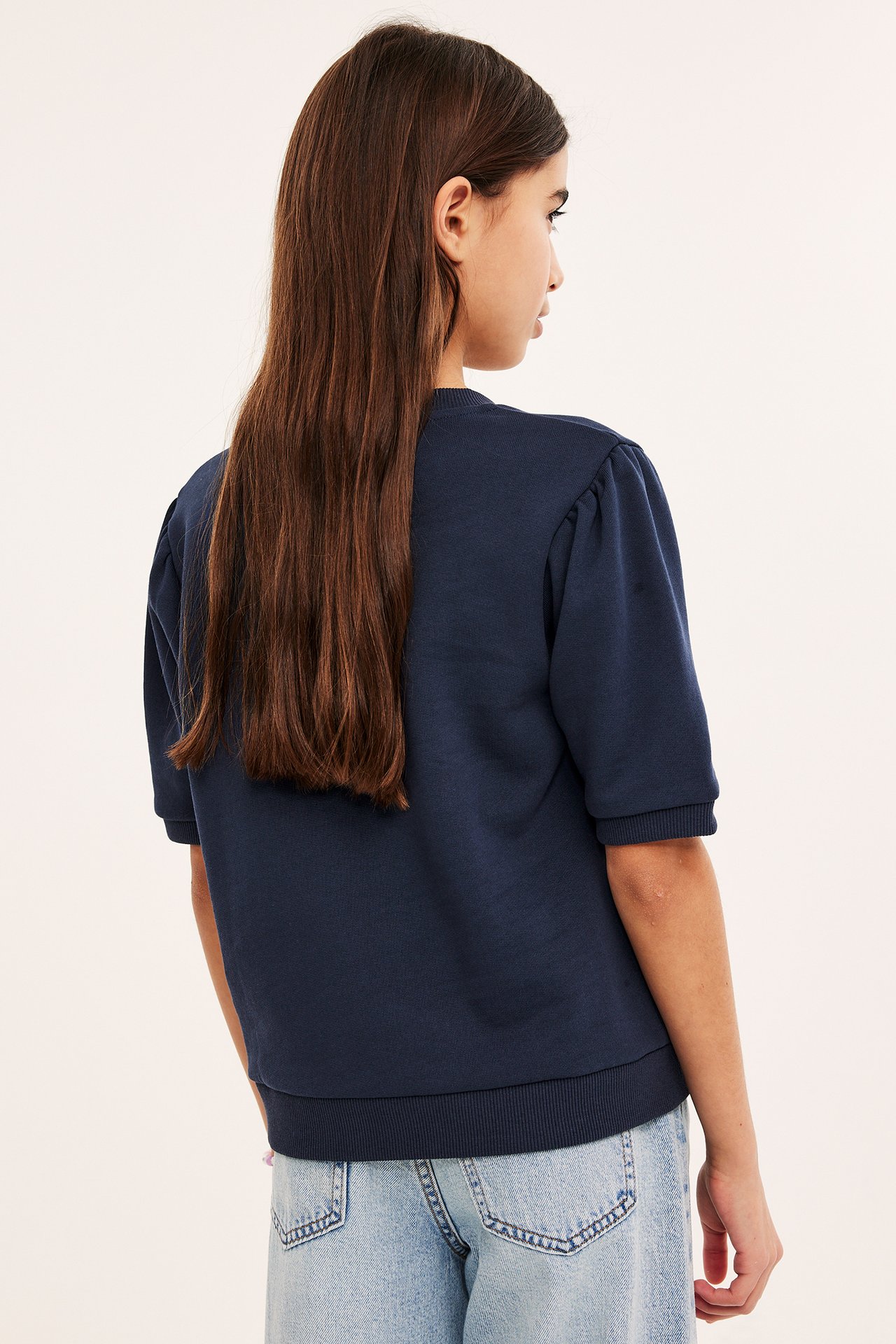 Rygg/sidovy av ett barn i en mörkblå kortärmad bomullssweatshirt med puffärmar och ljusblå jeans.