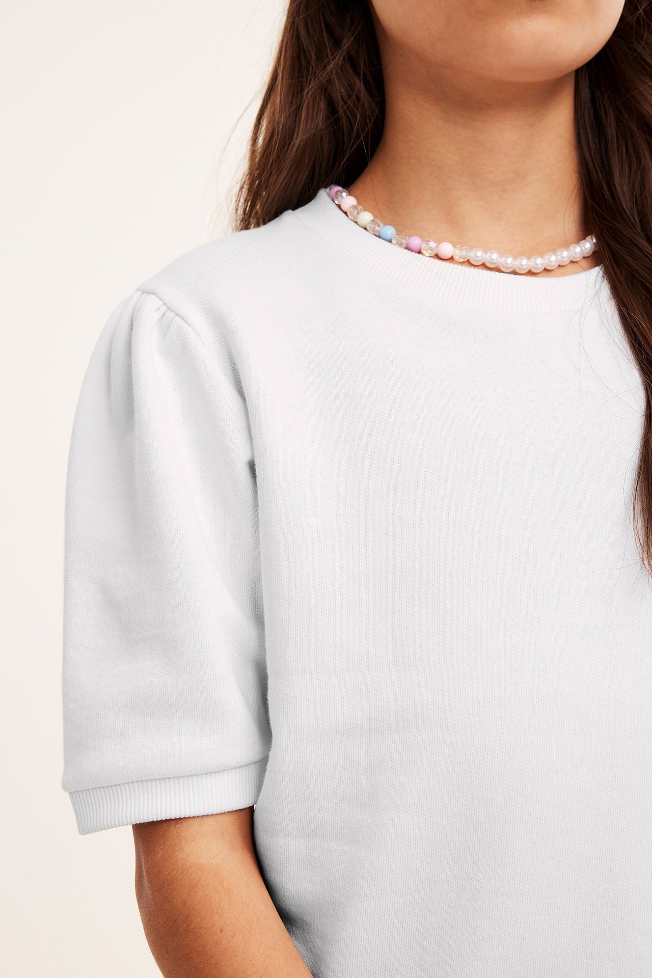 Lähikuva lapsesta valkoisessa lyhythihaisessa collegepaidassa, jossa pussihihat ja helmikaulakoru.