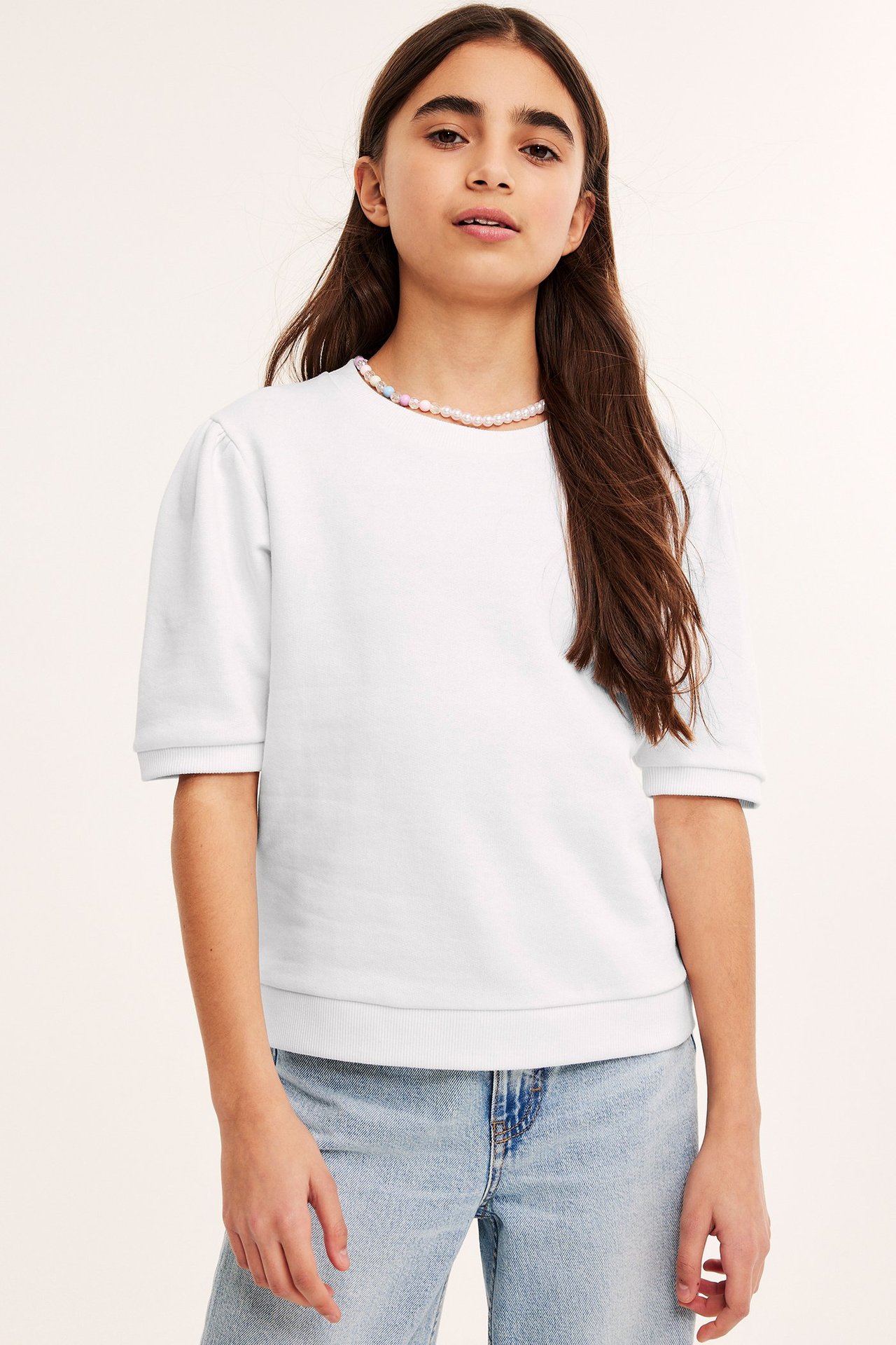 Edestäpäin: lapsi valkoisessa pussihihaisessa puserossa, vaaleansiniset farkut ja monivärinen helmikaulakoru.