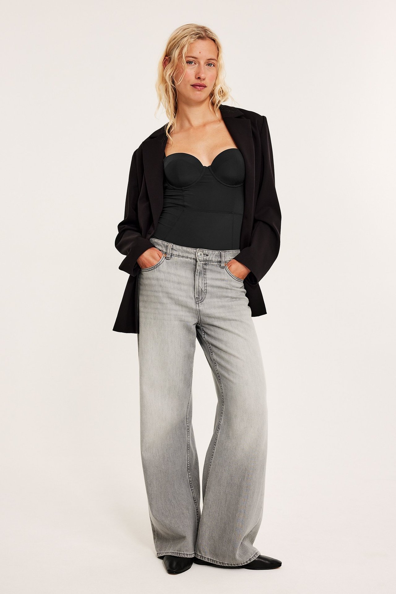 Forfra: kvinne i svart skulpturerende body, åpen svart blazer og lysegrå vide jeans.
