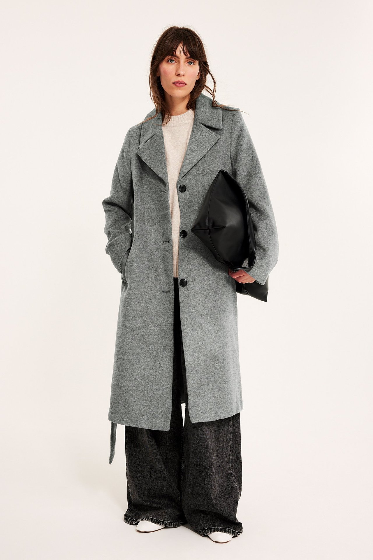 Edestä: Nainen harmaamelange-nappitakissa, beigessä neuleessa, mustissa farkuissa, pitäen mustaa käsilaukkua.