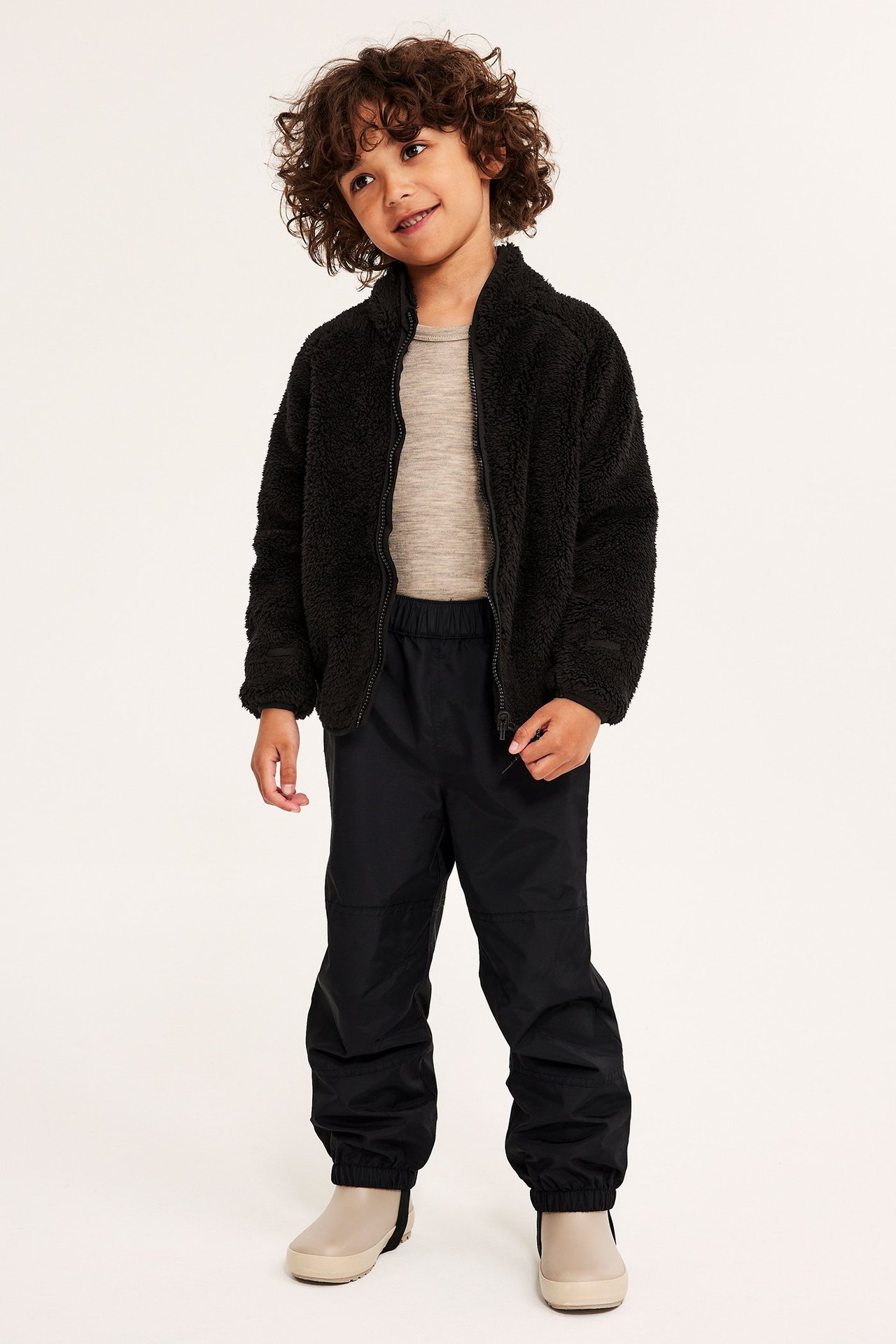 Edestäpäin, lapsi mustassa pilefleece-takissa, vaaleanbeigessä paidassa, mustissa housuissa ja beigessä saappaissa.