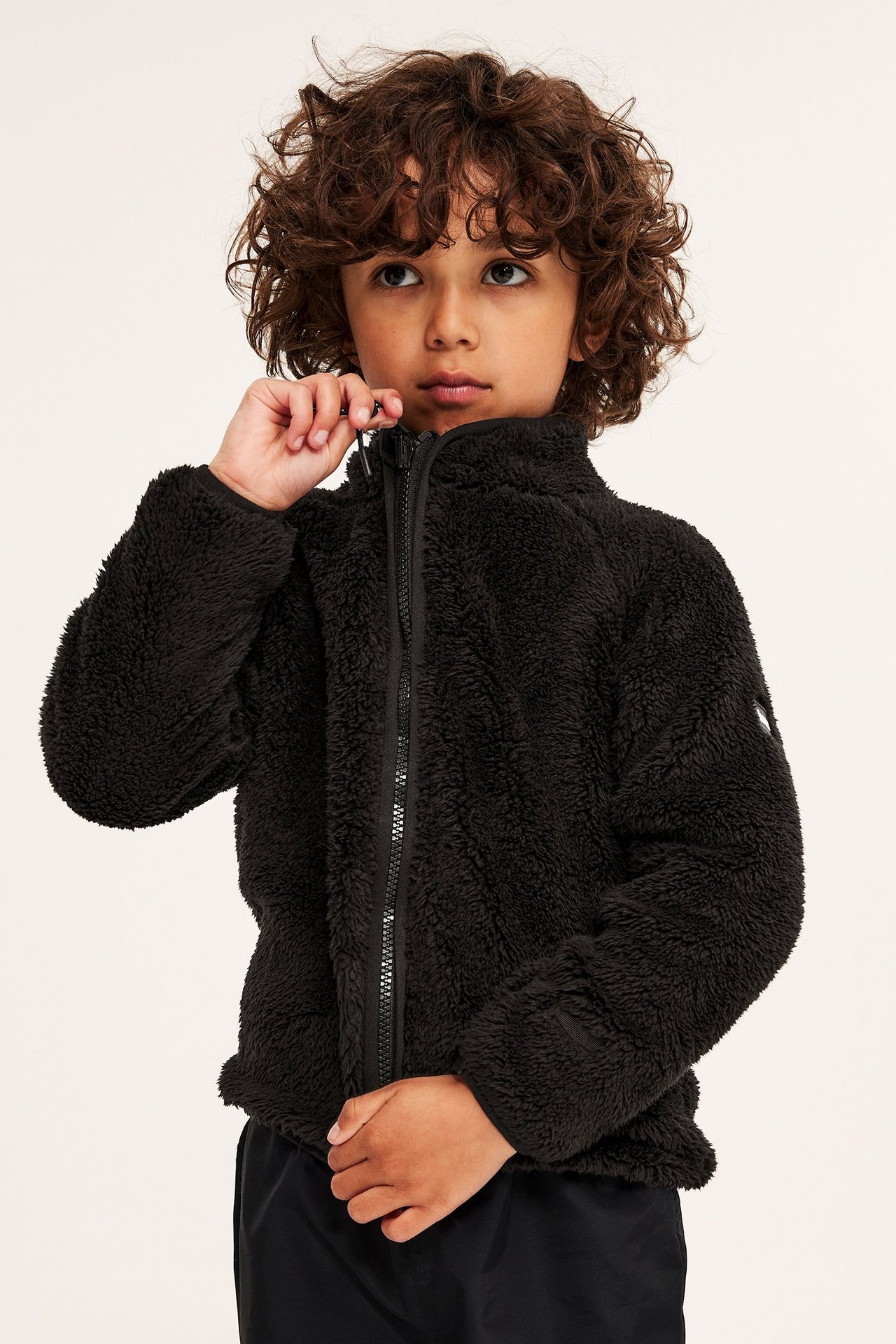 Framifrån, ett barn drar upp en svart pilefleecejacka med hög krage, bärs med svarta byxor.