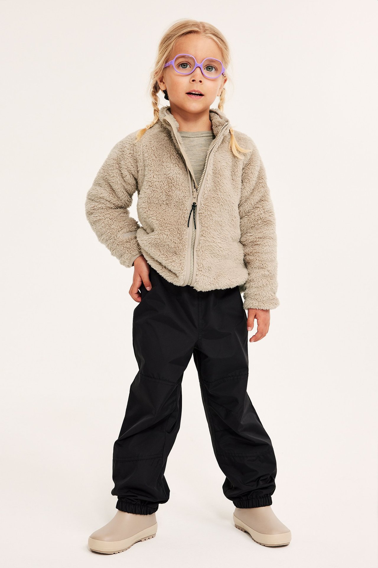 Edestä: lapsi beigessä pilefleecetakissa, harmaassa paidassa, mustissa housuissa, beigenvärisissä saappaissa, violeteissa laseissa.