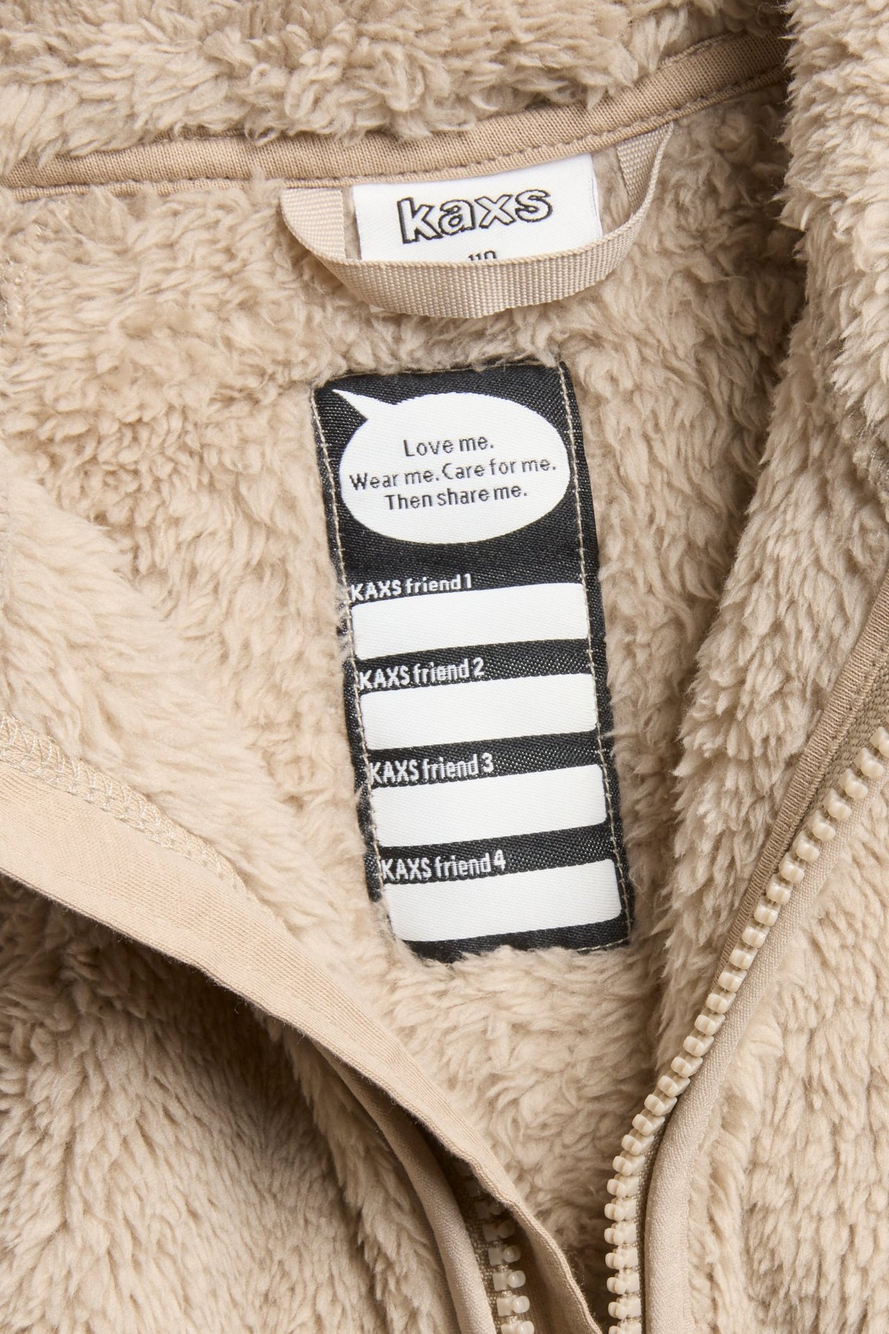 Zbliżenie beżowej kurtki polarowej od wewnątrz, z metką Kaxs, etykietą "Love me" i zamkiem błyskawicznym.