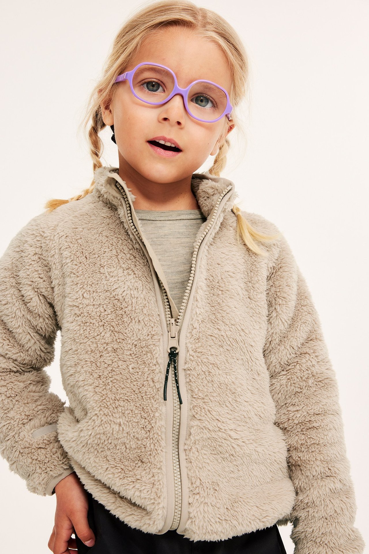 Frontvy av ett barn i beige pilefleecejacka med dragkedja över grå topp, med lila glasögon.