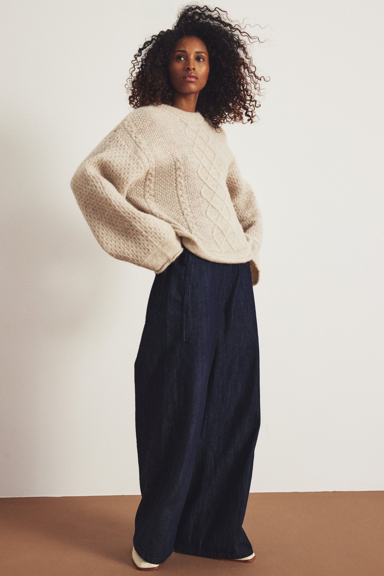Frontvy av kvinna i mörkblå vida denimjeans och en beige kabelstickad tröja.