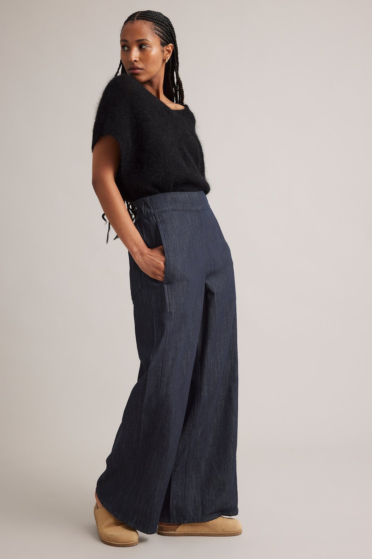 Framifrån: kvinna i mörk denim vida jeans med fickor, svart luddig topp och ljusbruna träskor.