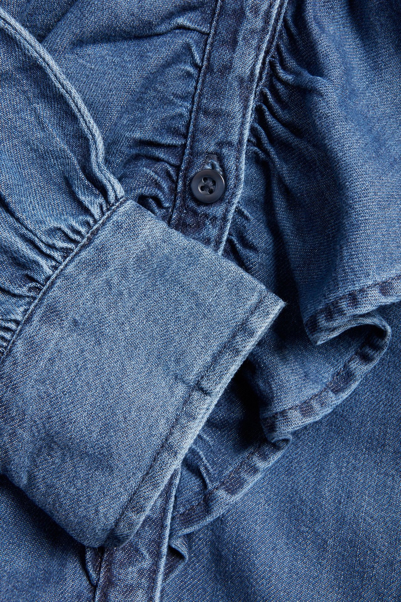 Zbliżenie niebieskiej bluzki jeansowej z zapinanym mankietem, plisą guzikową i falbanami.