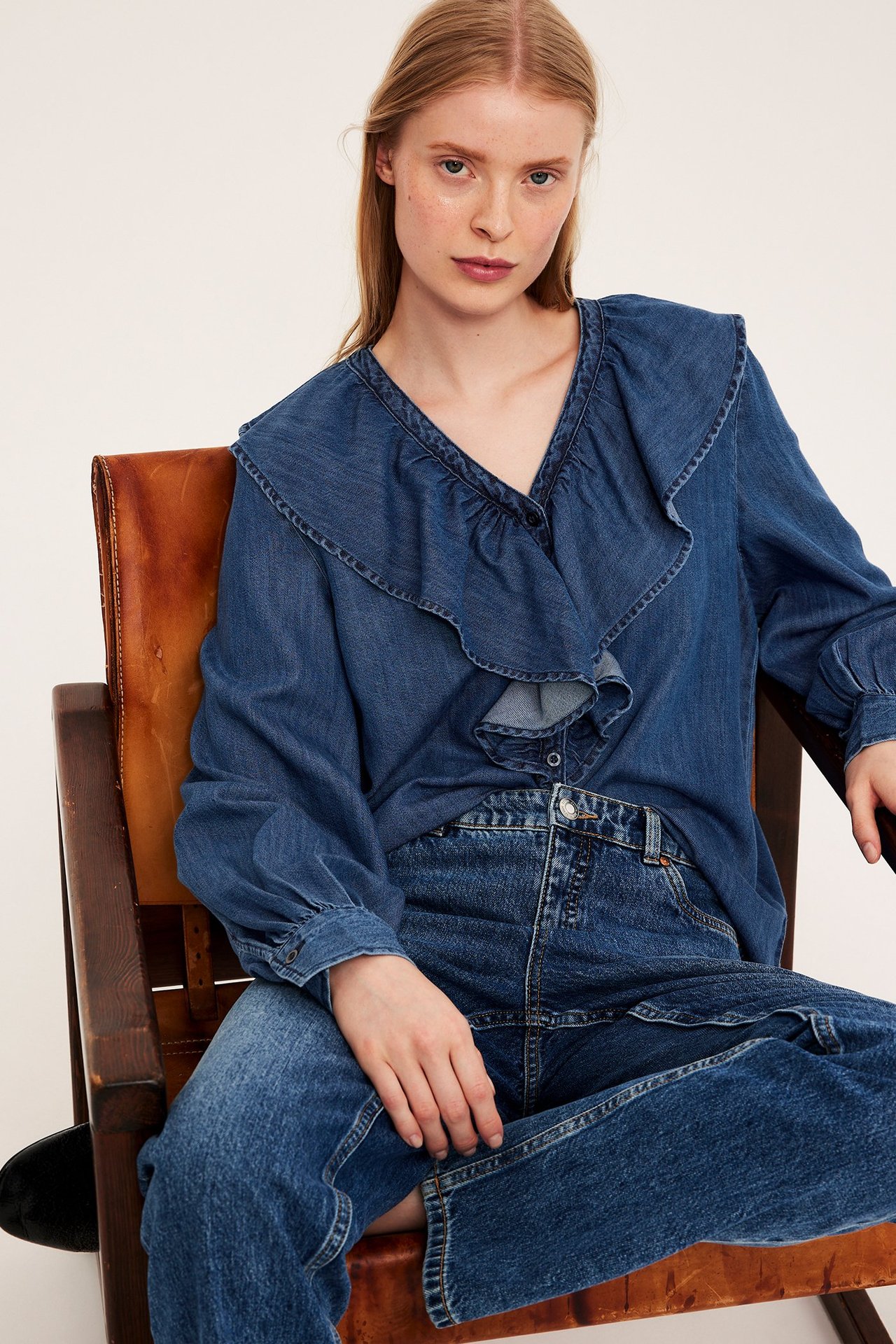 Widok z przodu kobiety w niebieskiej bluzce jeansowej z dekoltem V, falbanami i guzikami, oraz niebieskich jeansach.