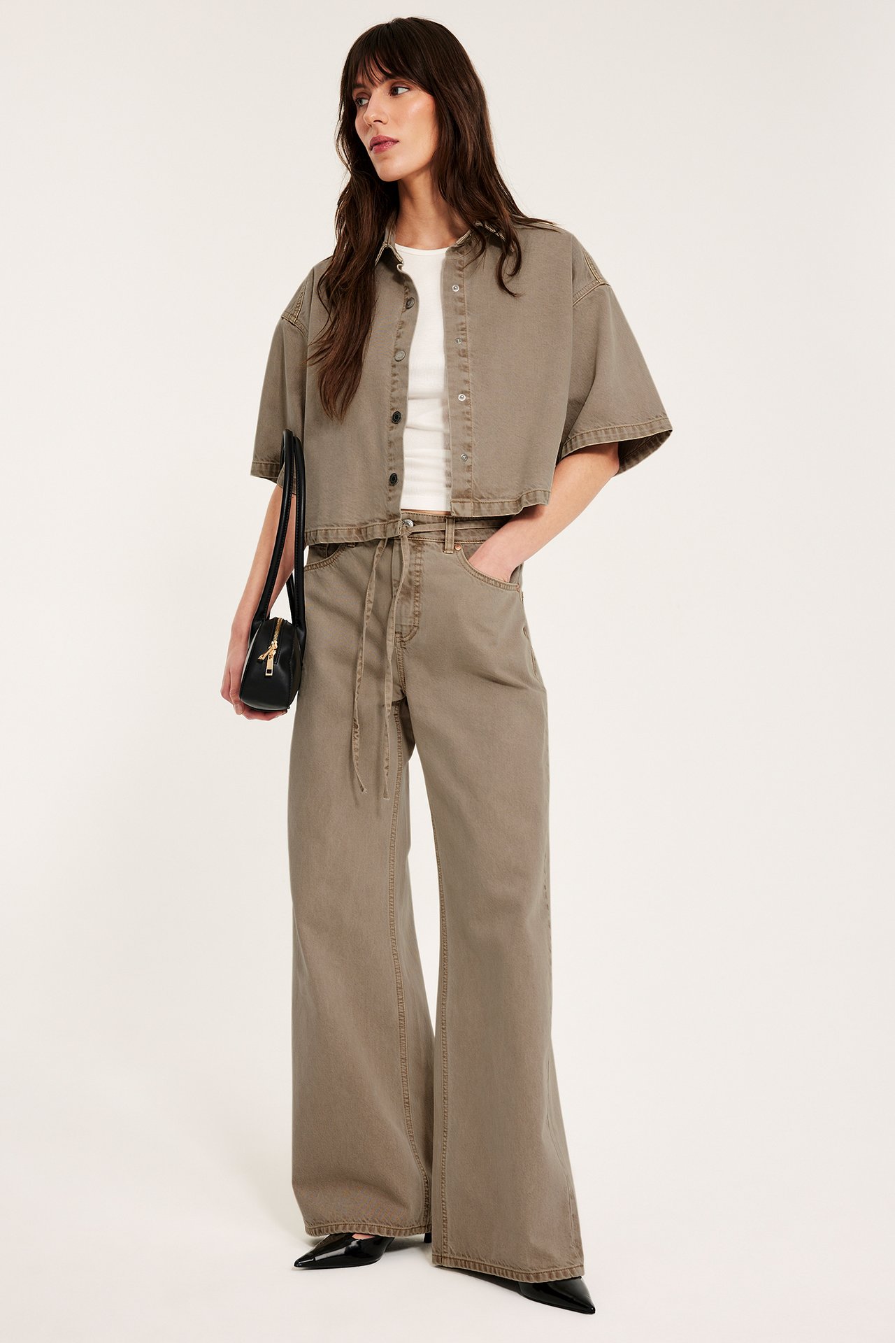 Frontvy av kvinne i brun kortermet twillskjorte over hvit topp, matchende vide bukser og svart håndveske.