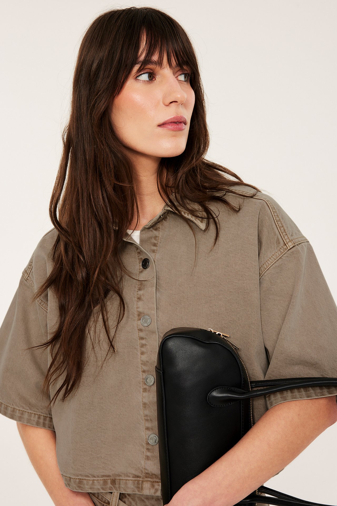 Nærbilde av en kvinne i en kortermet brun twillskjorte med dongeriknapper, som holder en svart håndveske.
