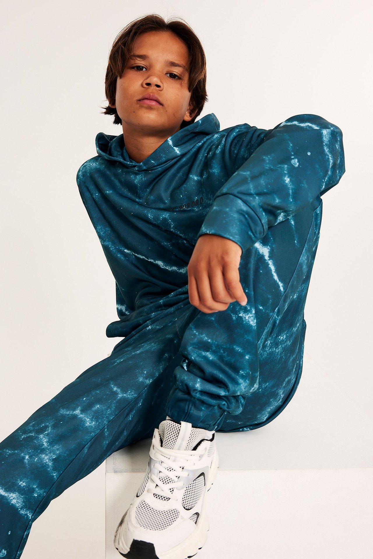 Widok z przodu dziecka w zielonej wzorzystej bluzie z kapturem, pasujących spodniach i białych sneakersach.