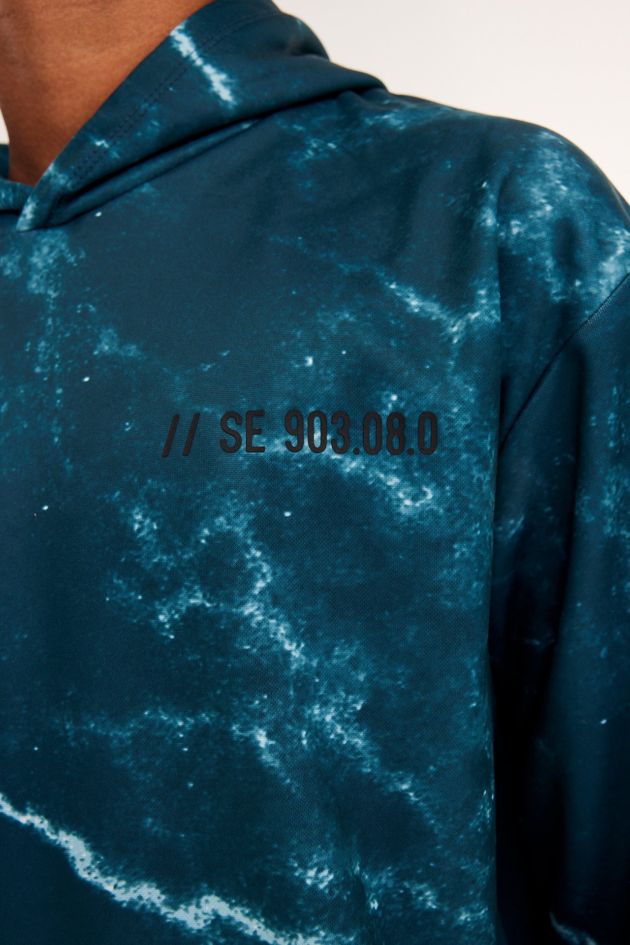 Närbild framifrån på ett barn i mörkgrön mönstrad polyesterhoodie med svart texttryck.