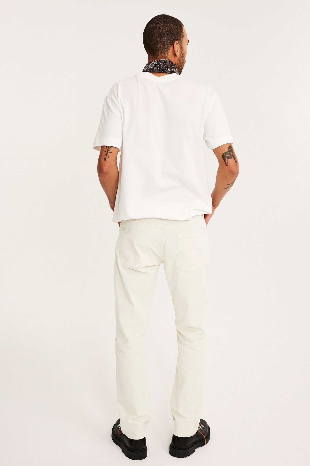 Bakifrån, man i offwhite manchesterbyxor, vit t-shirt, svart mönstrad bandana och svarta skor.