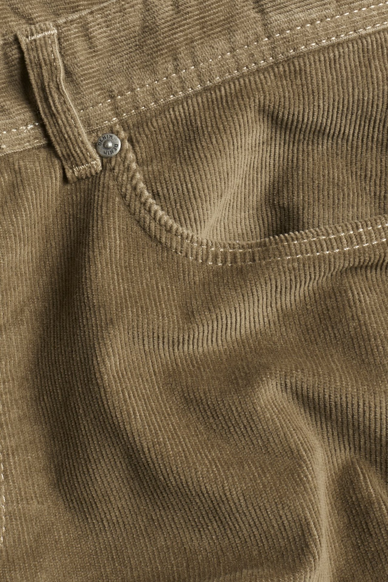 Zbliżenie brązowych sztruksowych spodni, ukazujące przednią kieszeń ze srebrnym guzikiem i białymi szwami.