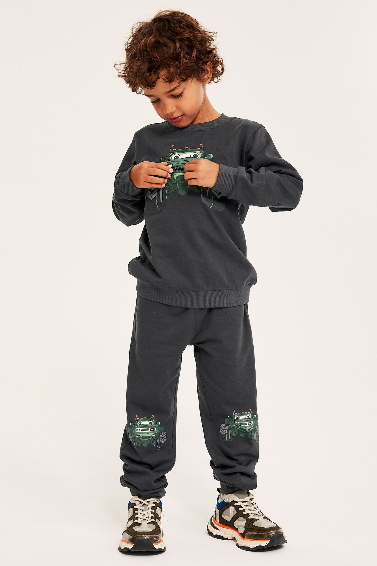 Frontbilde av et barn i mørkegrå bomullsgenser med monstertruck-trykk og matchende joggebukse.