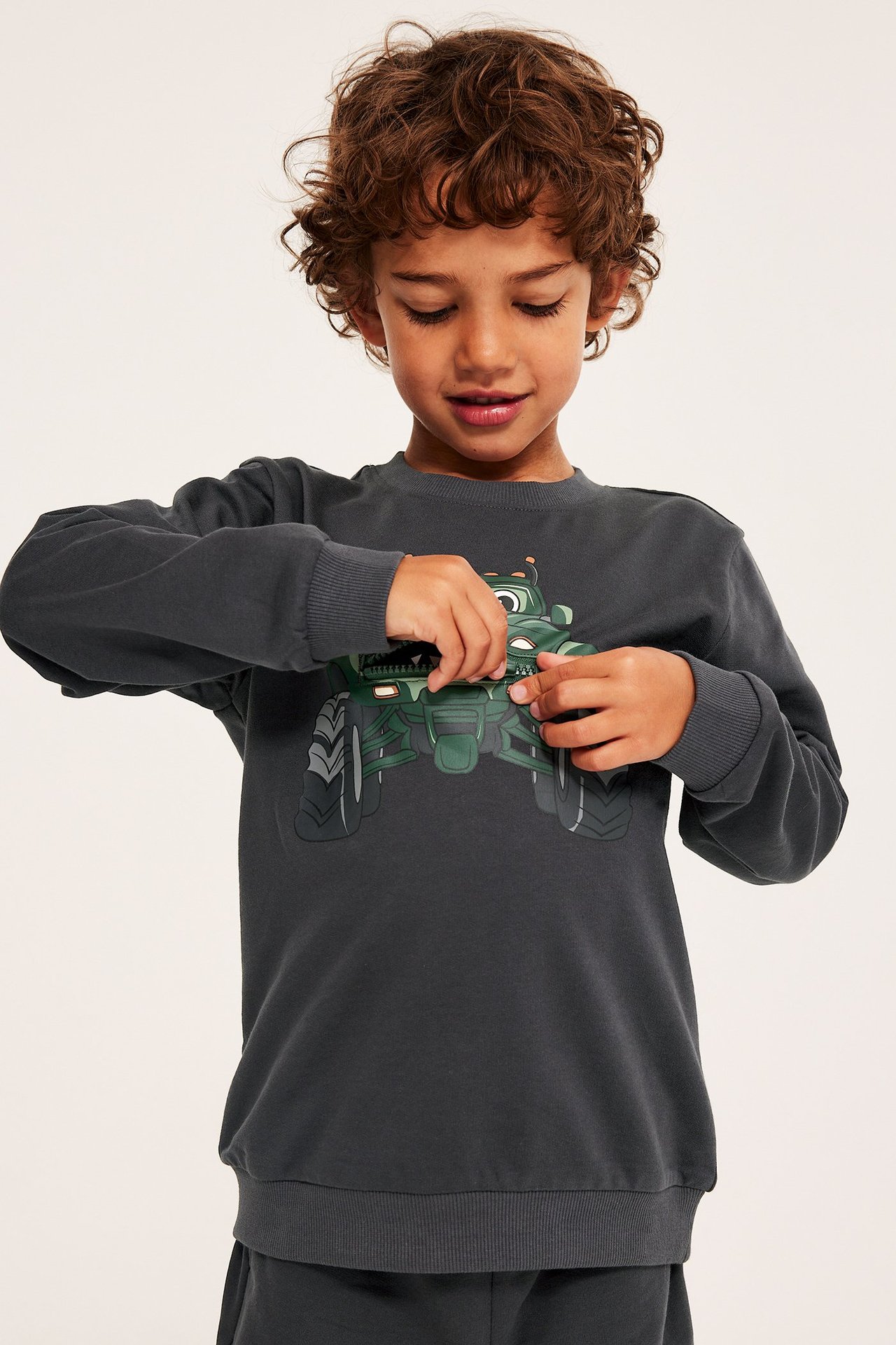 Widok z przodu dziecka w czarnej bawełnianej bluzie z nadrukiem monster trucka, bawiącego się zamkiem.