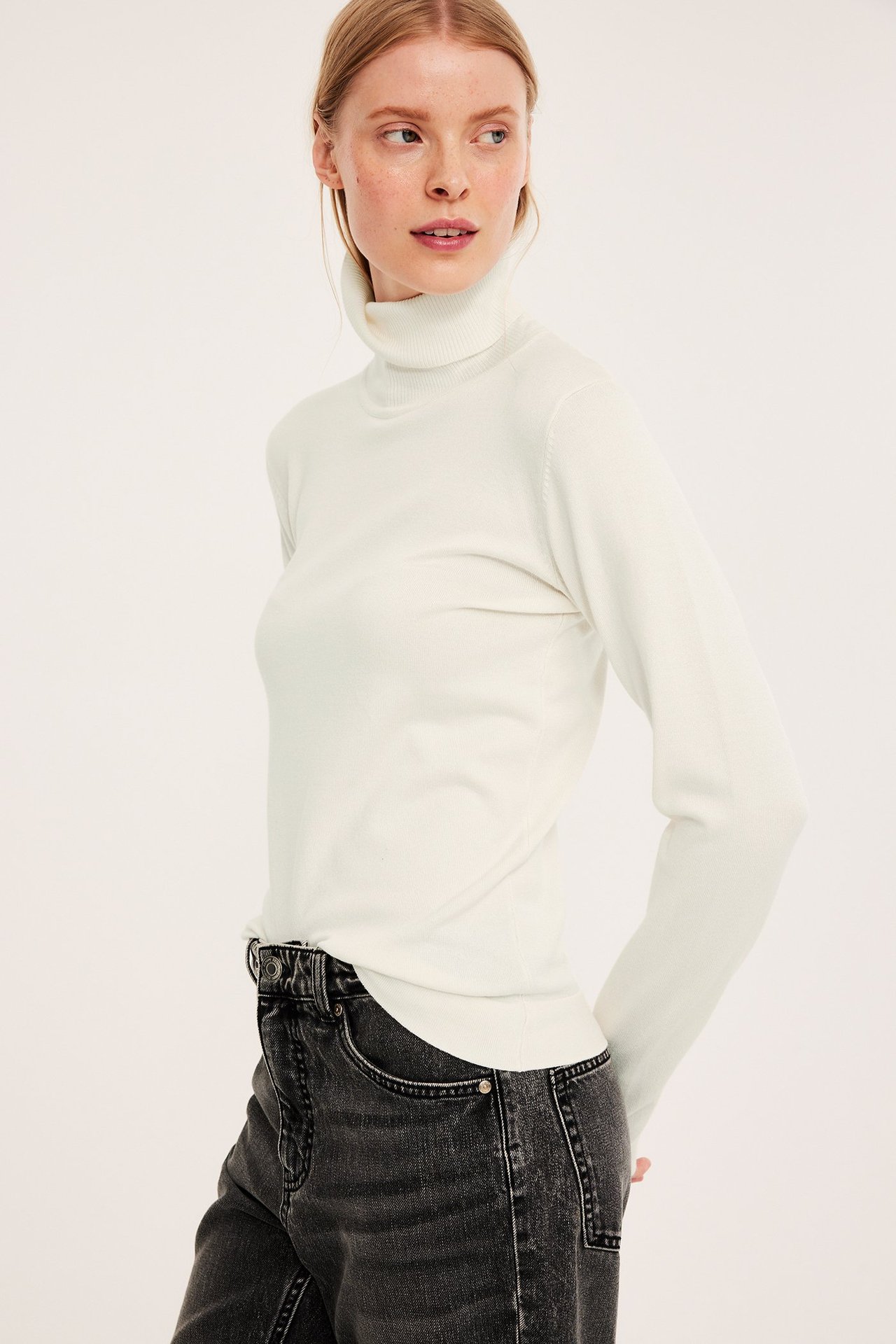 Frontbilde av kvinne i en smal, offwhite finstrikket høyhalset genser stukket ned i mørkegrå jeans.