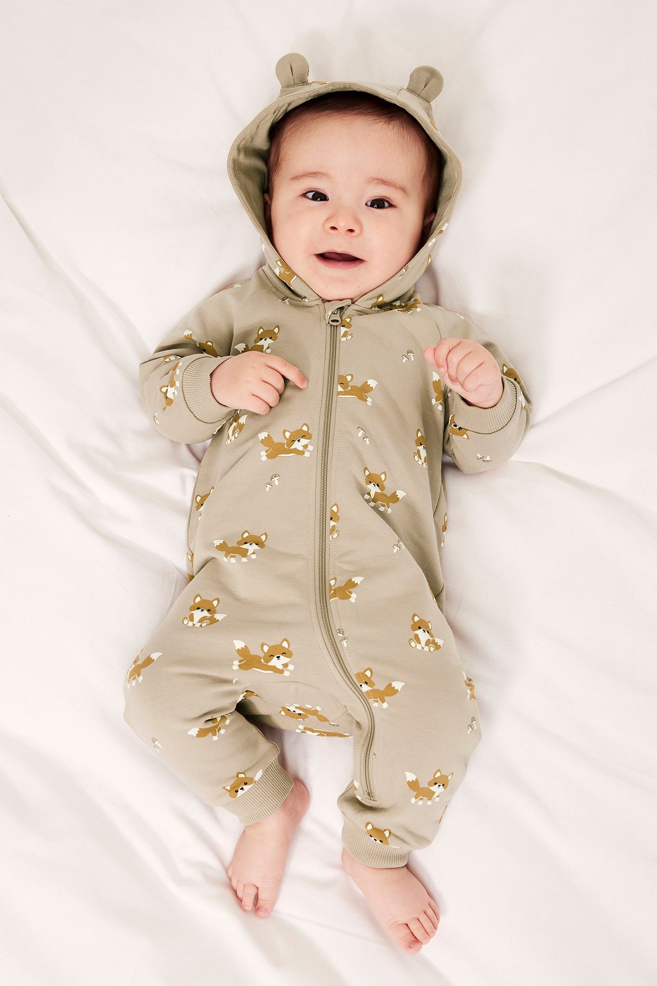 Ylhäältä päin: vauva beigessä kettukuvioisessa hupullisessa haalarissa, jossa vetoketju ja korvat.