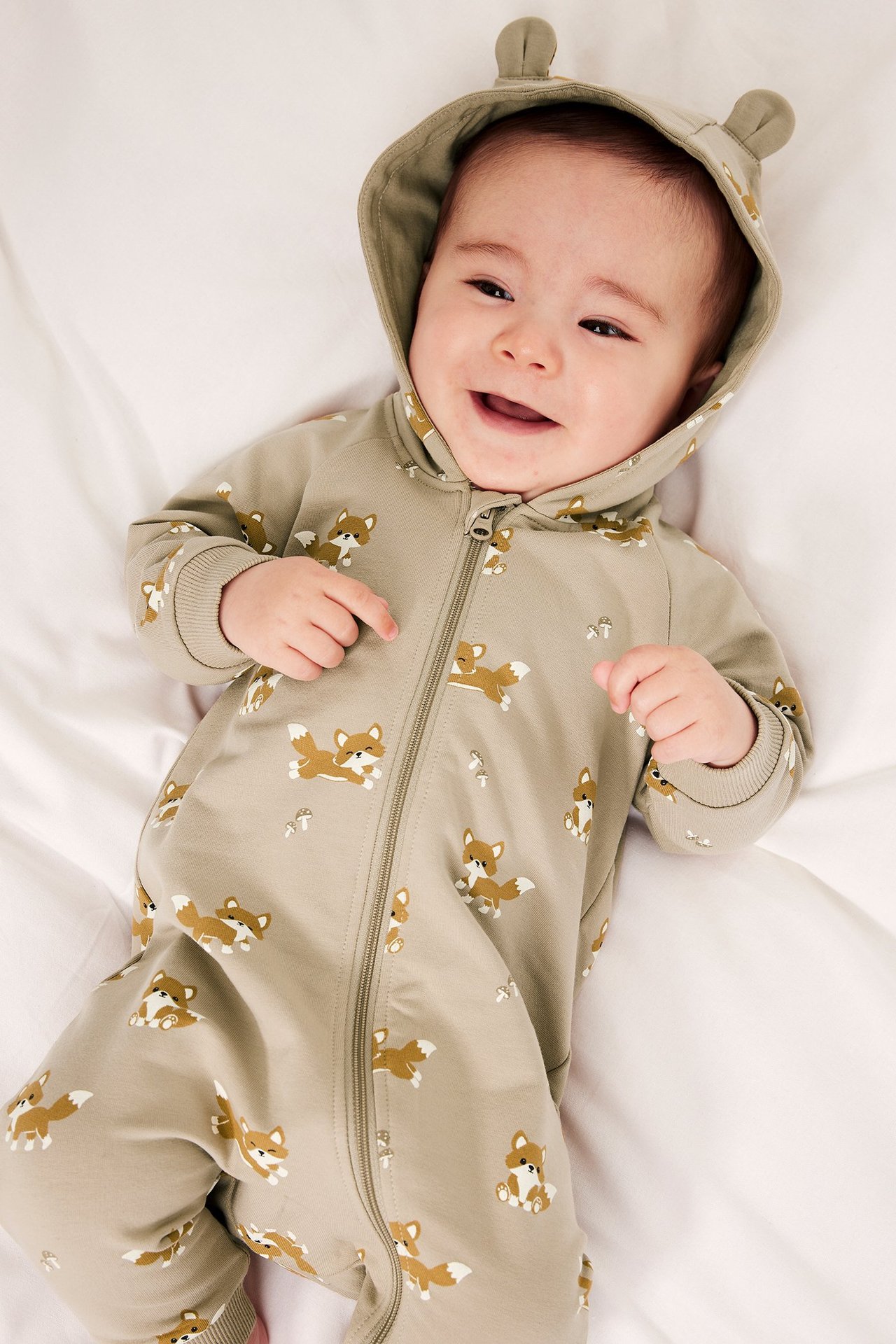 Leende baby i beige räv-mönstrad jumpsuit med dragkedja fram och huva med öron.