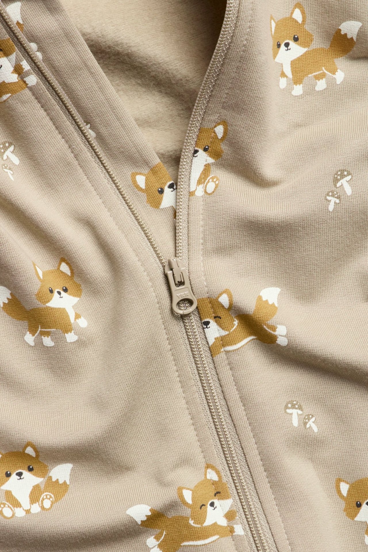 Zbliżenie beżowego pajacyka niemowlęcego z uroczym wzorem w lisy i grzyby, z zamkiem błyskawicznym z przodu.