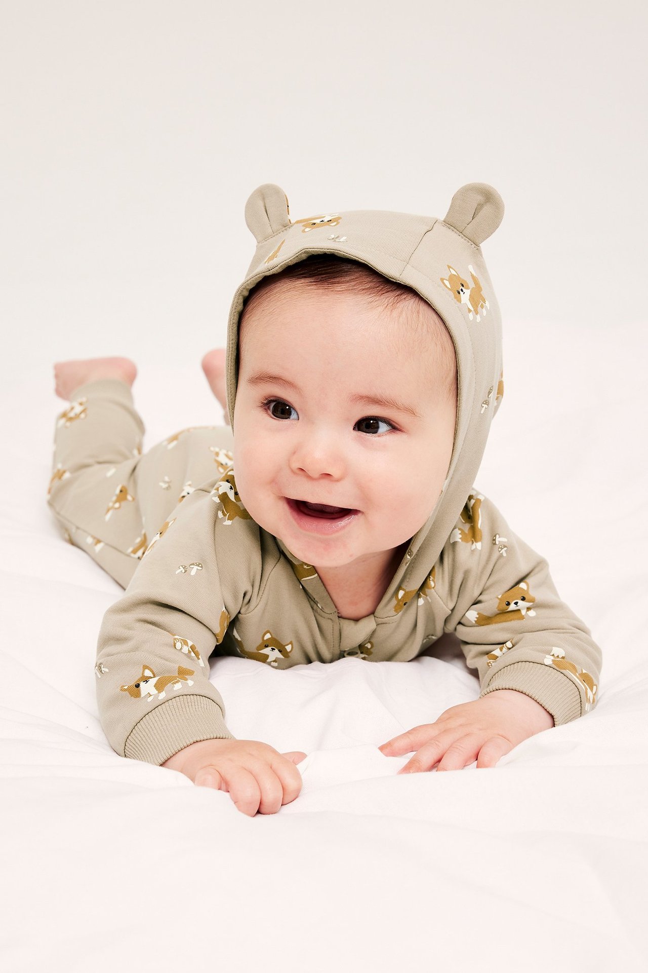 Edestä kuvattu vauva yllään beige kuvioitu haalari, jossa on korvallinen huppu ja vetoketju.