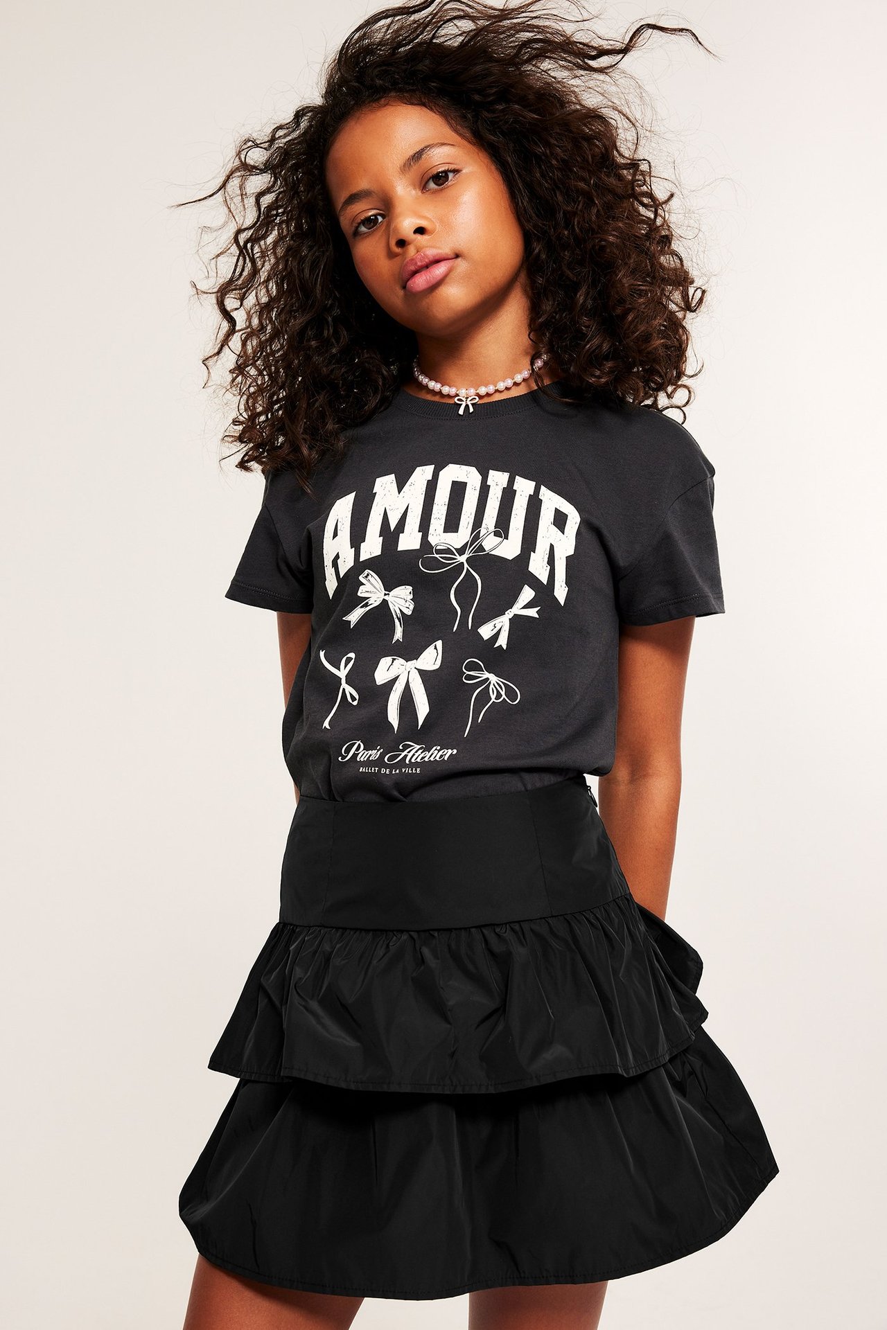 Framsida av ett barn i svart volangkjol med volanger, mörkgrå AMOUR t-shirt och rosa pärlhalsband.