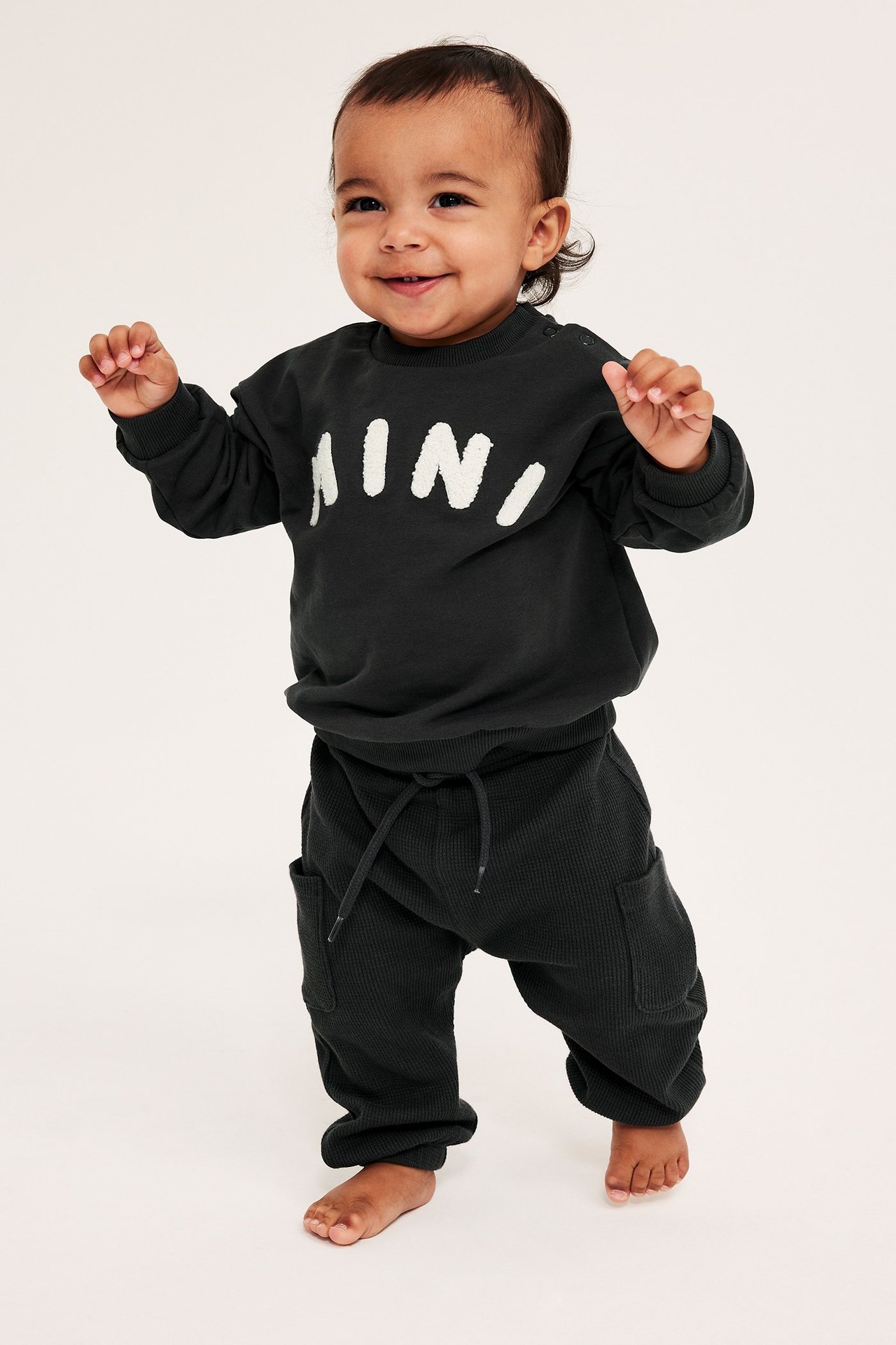 Edestäpäin vauva mustassa collegepaidassa, jossa valkoinen 'MINI'-applikaatio, ja mustissa vohvelihousuissa.