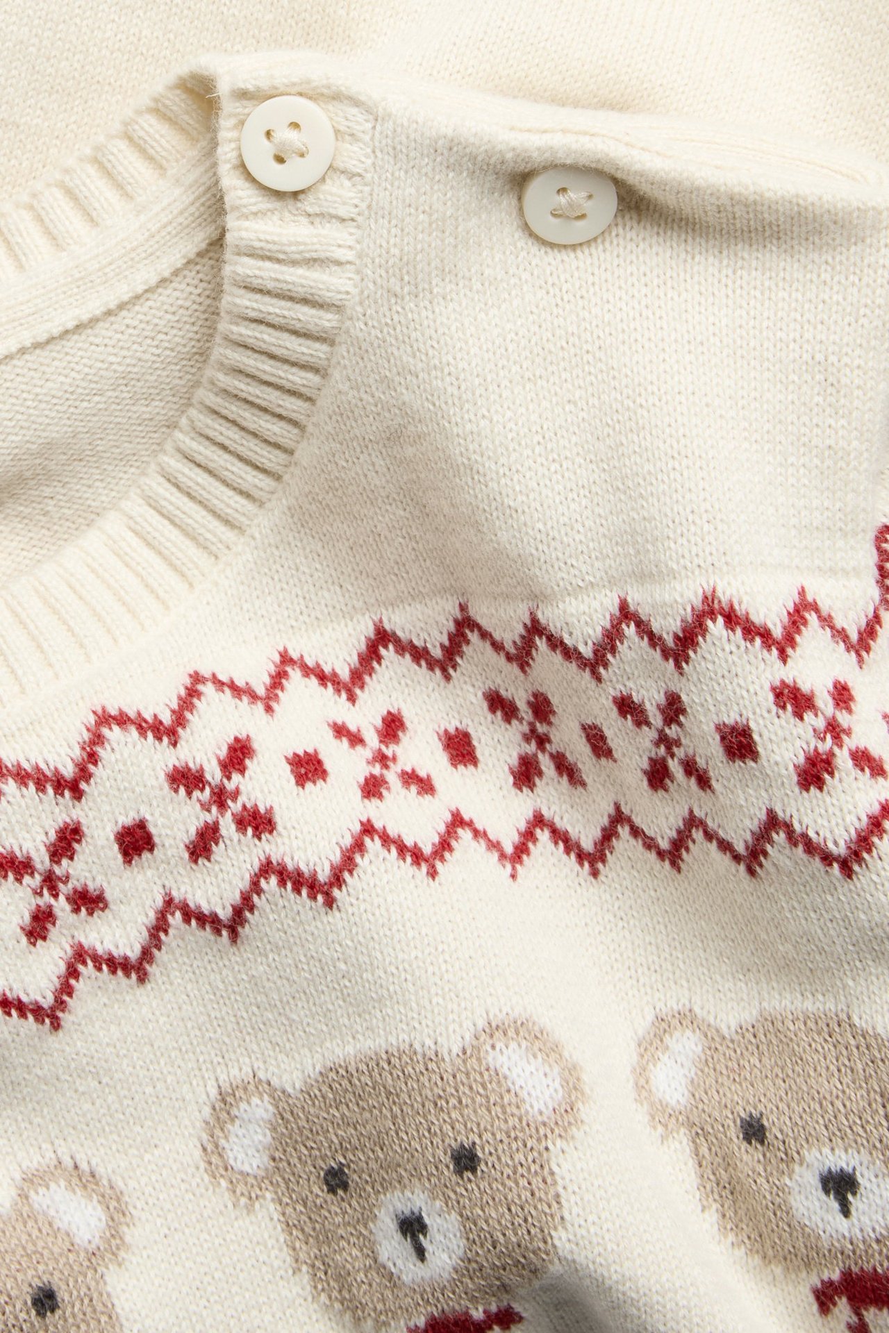 Lähikuva vauvan vaaleanbeigestä puuvillaneuleesta, jossa punainen jacquard-kuvio ja karhuja, napit olkapäällä.
