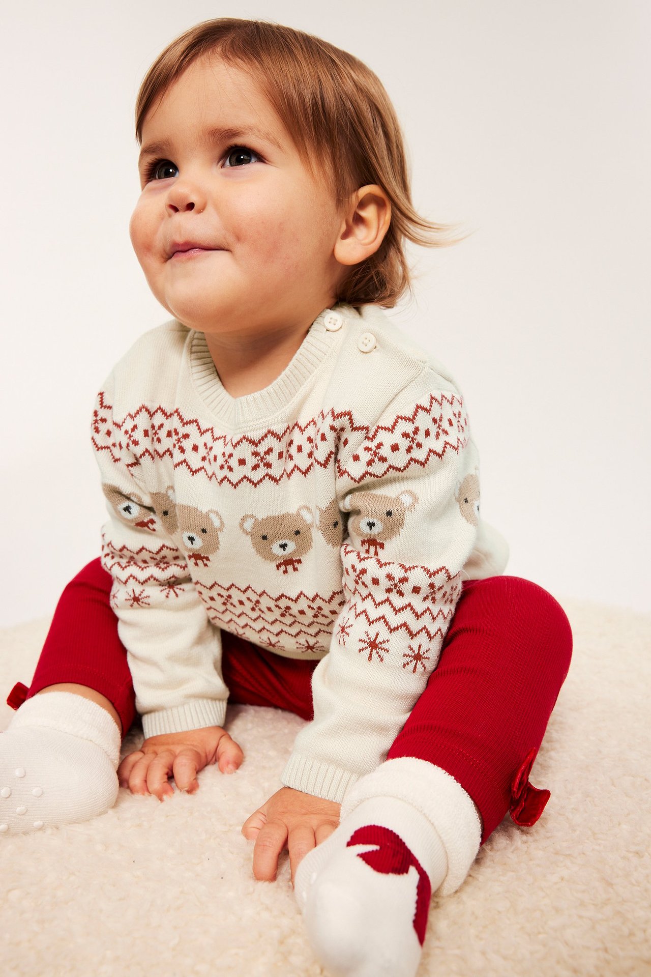 Baby i lys beige julegenser med rød mønster, røde bukser, sitter på gulvet.