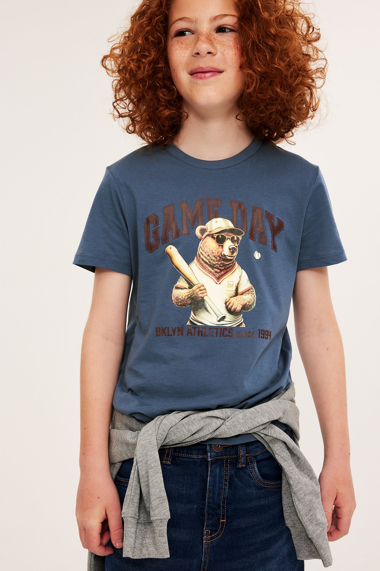 Frontvy: Ett barn bär en blå bomulls-t-shirt med basebollbjörntryck, mörkblå jeans och en grå sweatshirt.