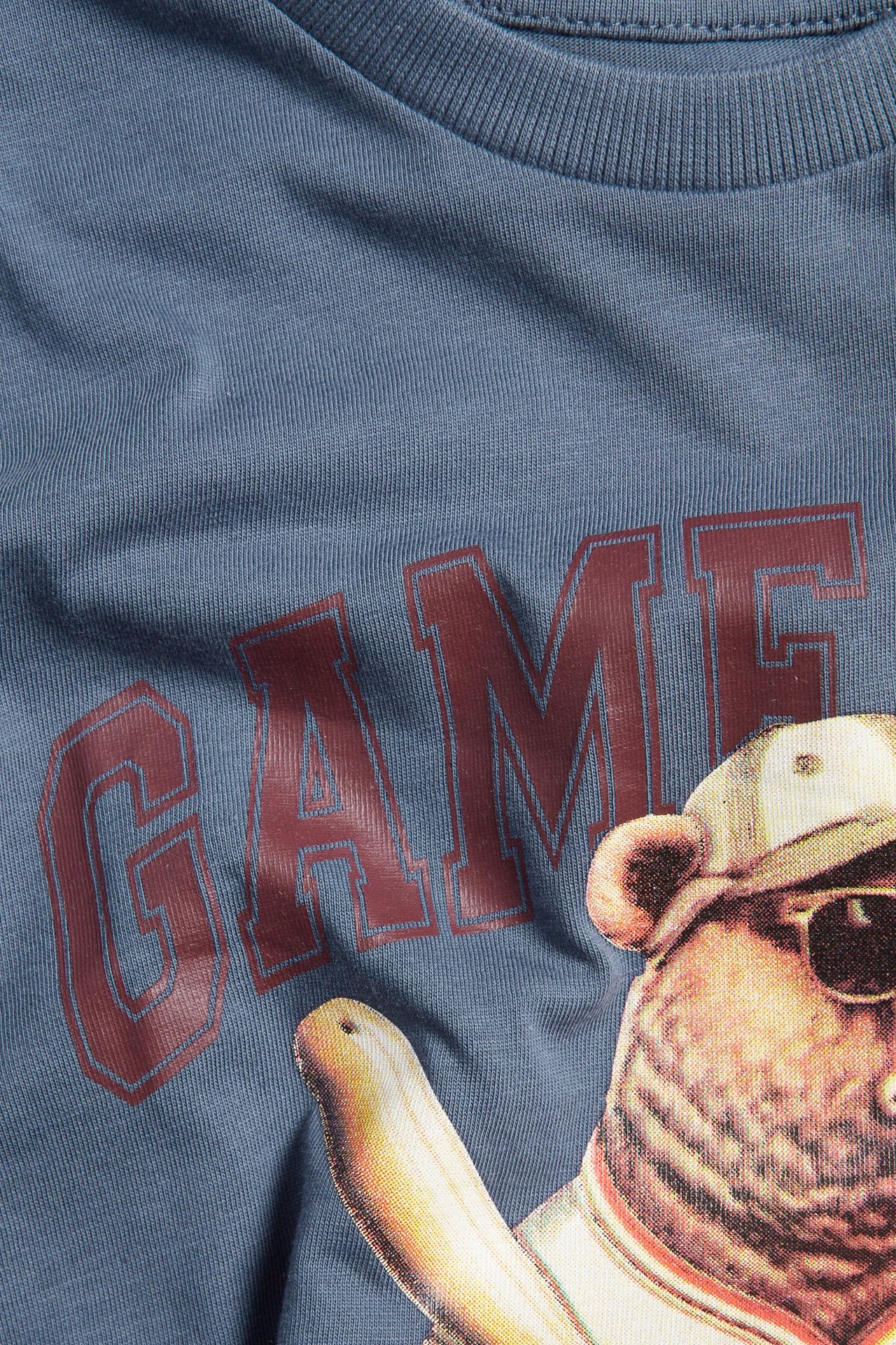 Zbliżenie na niebieski bawełniany T-shirt dziecięcy z prążkowanym dekoltem i nadrukiem misia z napisem „GAME”.